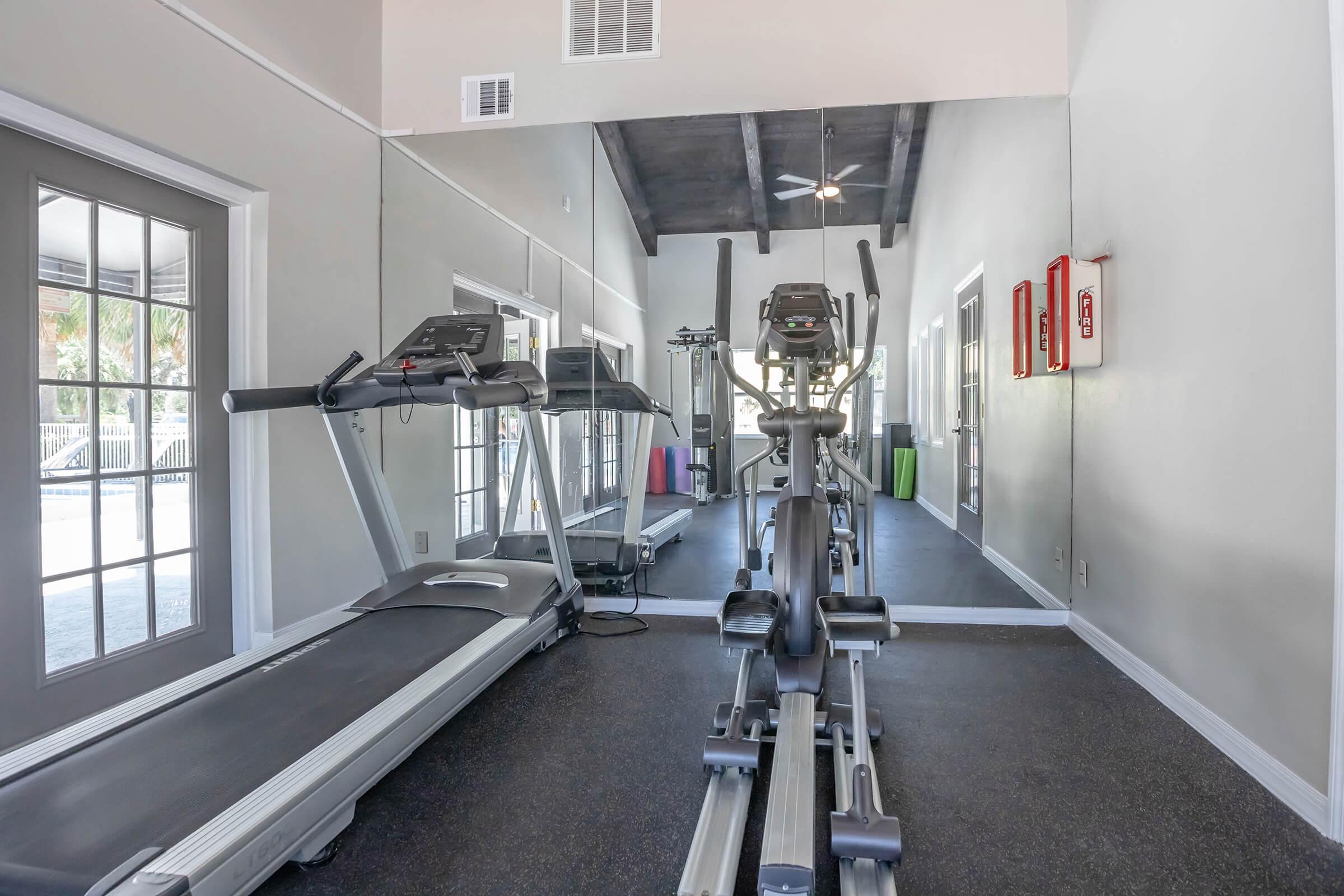 A modern gym interior featuring a treadmill and an elliptical machine, both positioned facing a large mirror. The walls are painted in light colors, and the space has large windows allowing natural light. An emergency equipment display is visible, adding safety features to the environment.