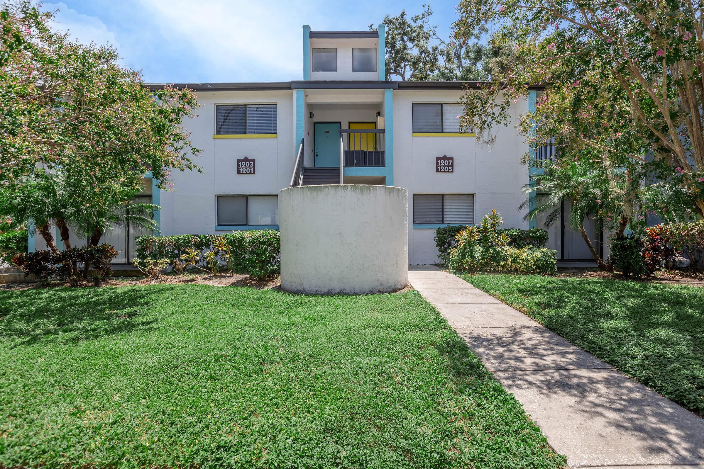 A two-story apartment building with light-colored walls and teal accents, featuring a round concrete planter in front. The entrance has a porch with colorful doors, and there are grassy areas and shrubs surrounding the walkway leading to the building. Bright blue sky visible in the background.