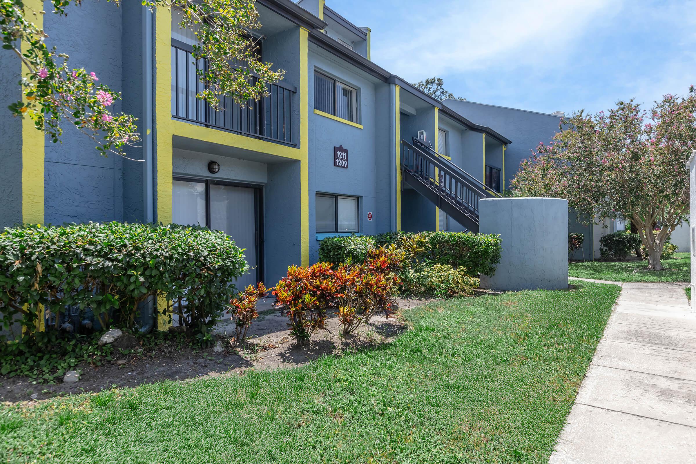 Exterior view of a two-story apartment building featuring blue and yellow accents. Lush green landscaping with flowering shrubs and neatly trimmed grass is visible in the foreground. A concrete pathway leads up to the entrance, and a balcony with railings is seen on the upper level. Clear blue sky in the background.