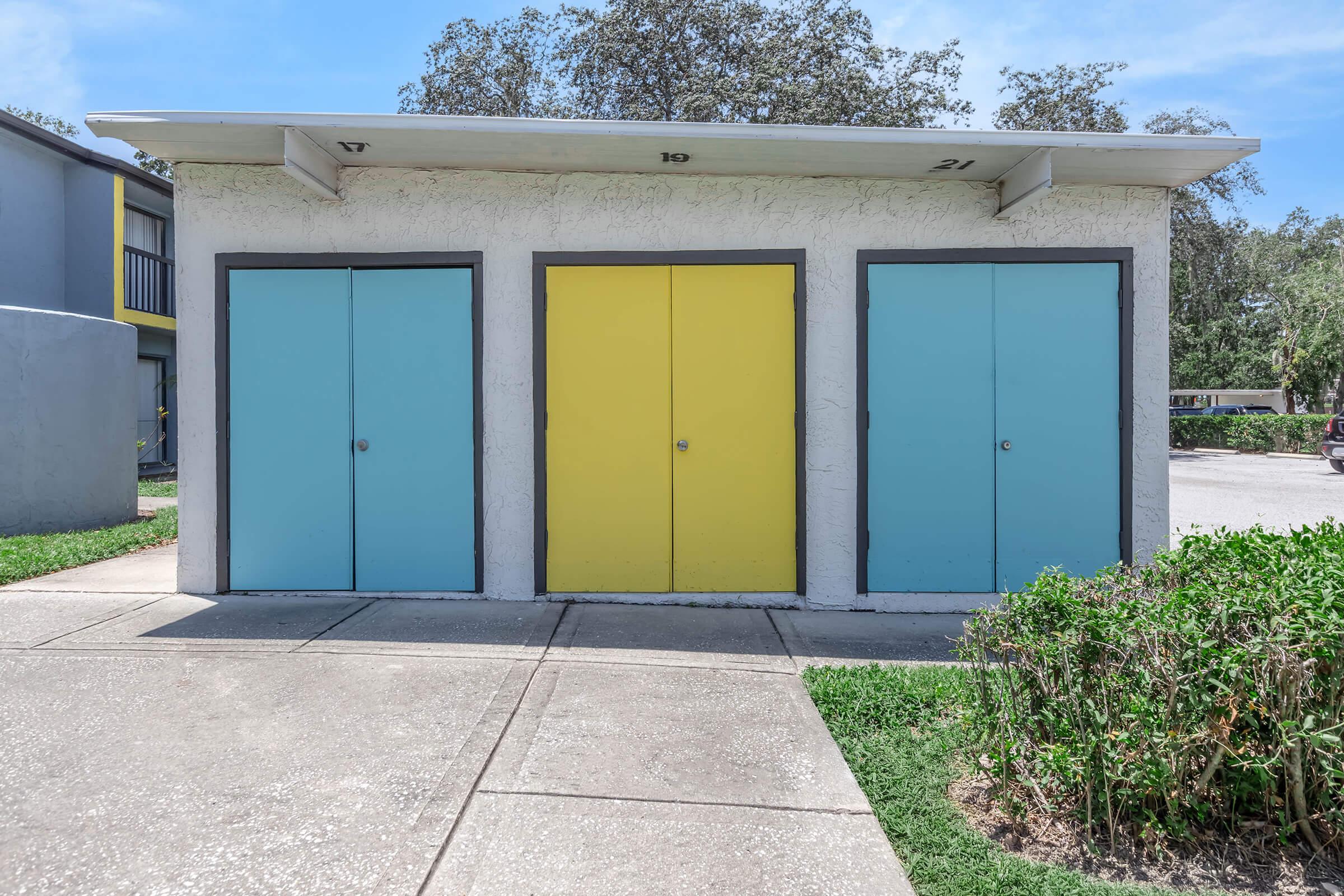 Three colorful storage doors arranged in a row against a light-colored wall. The doors are in shades of blue and yellow, with the blue doors on the left and right and a bright yellow door in the center. The building is surrounded by well-maintained grass and shrubs, set in a sunny outdoor environment.