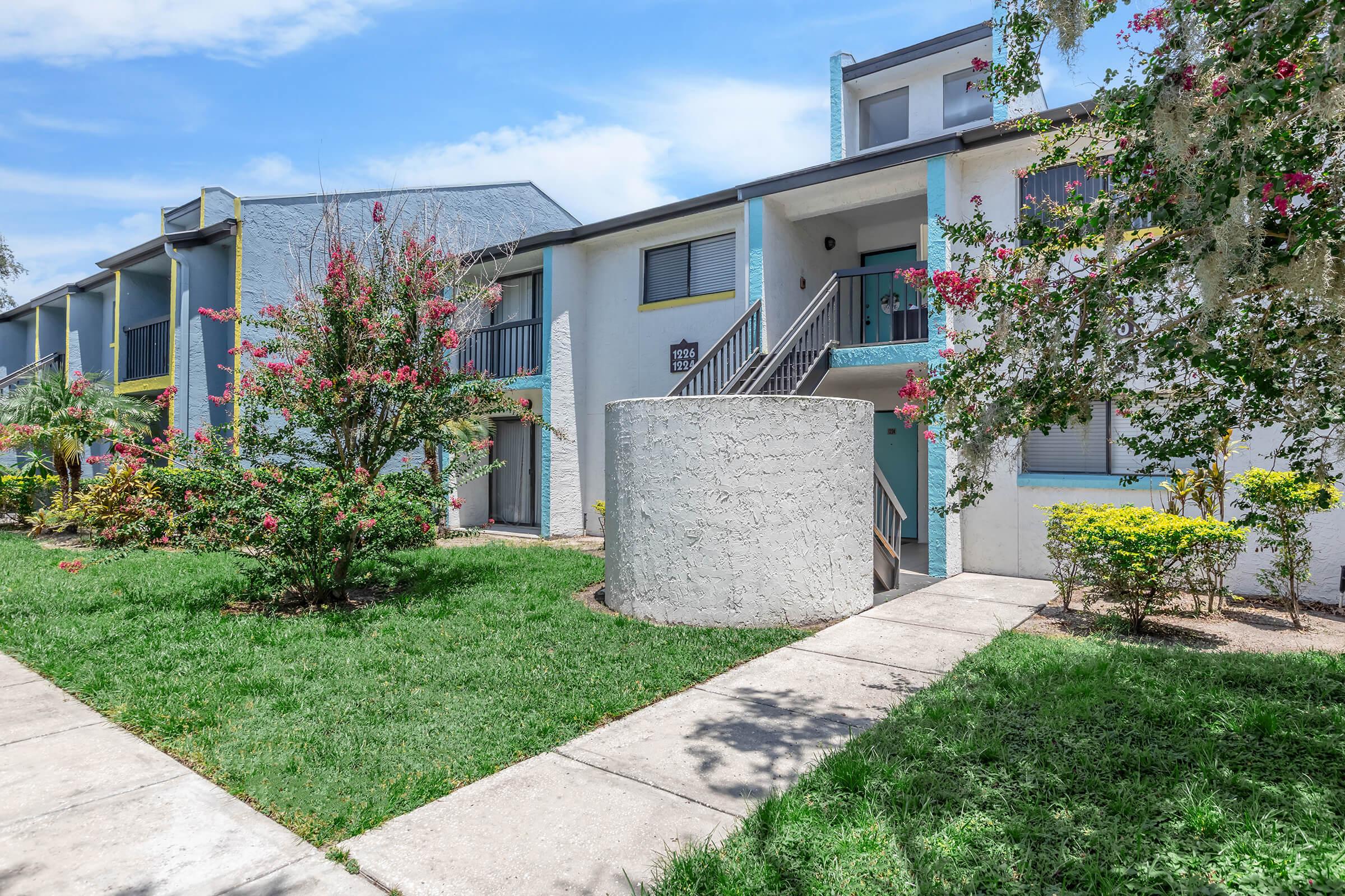 Exterior view of a multi-unit residential building featuring a light-colored facade with blue accents. A circular concrete structure is positioned in the foreground, surrounded by green grass and ornamental shrubs. Bright sunlight enhances the colors, and a clear blue sky is visible above.