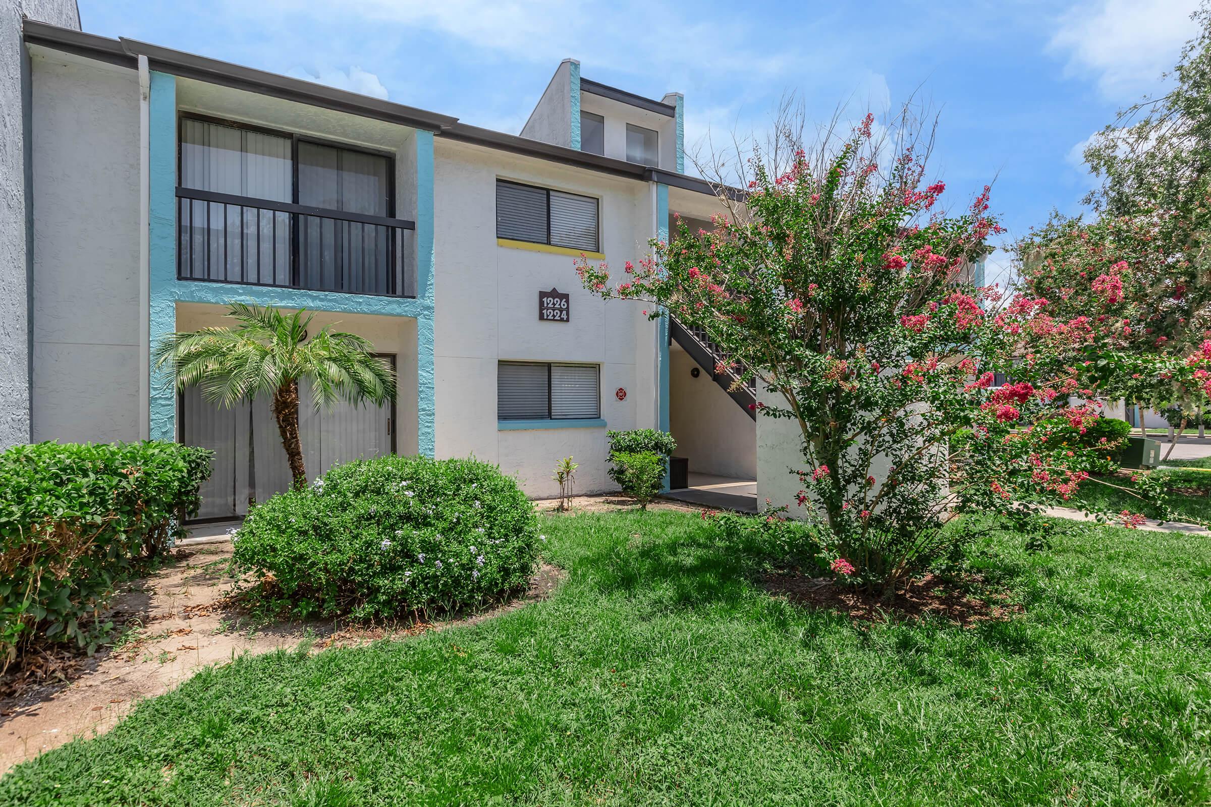 A two-story residential building with light blue and gray exterior. The entrance features dark railings and a small set of stairs. Lush green landscaping surrounds the building, including shrubs and a flowering plant. The sky is bright with a few clouds visible.
