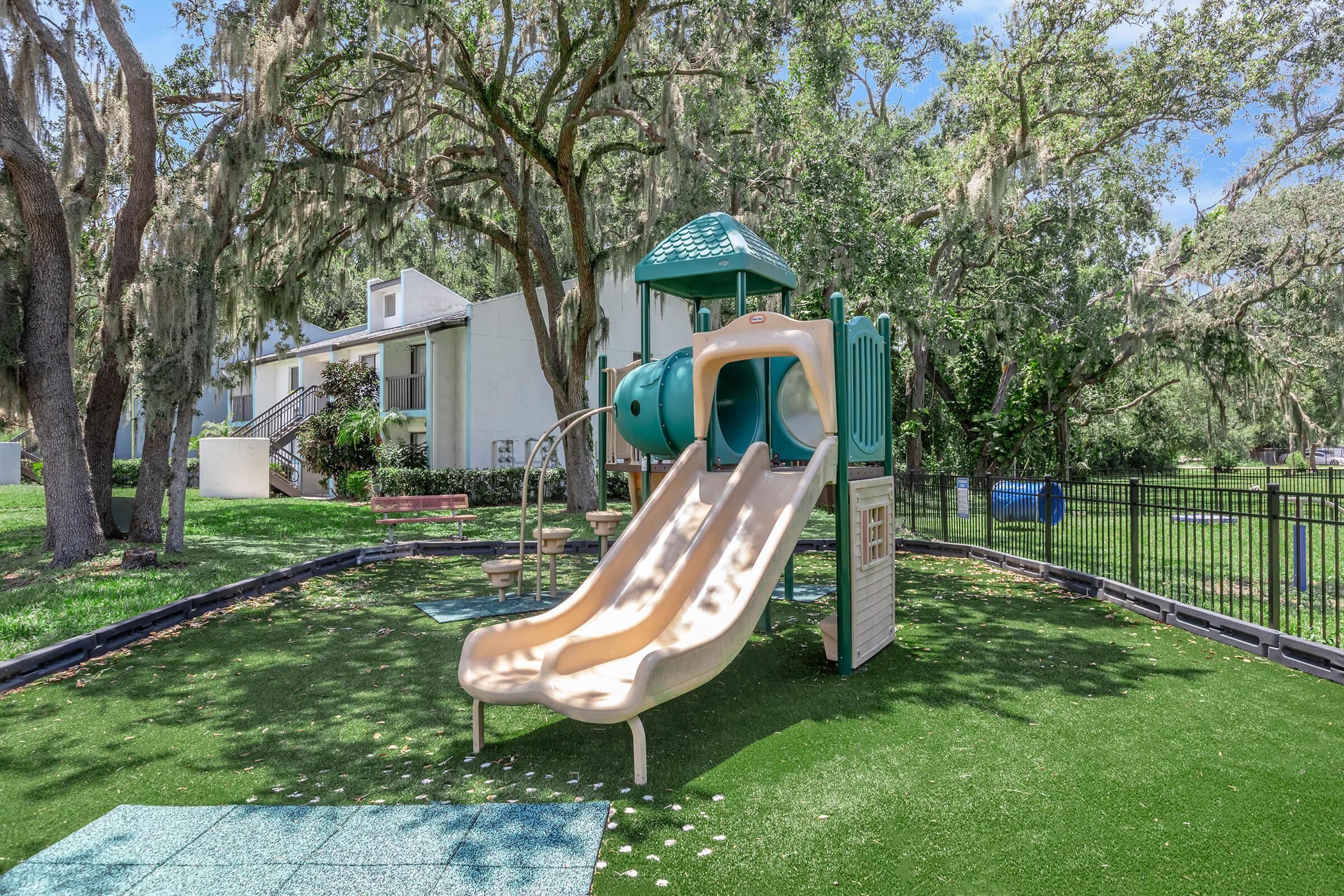 Playground with a green slide and climbing structure, surrounded by lush trees and grassy areas. A bench is visible in the background, and nearby buildings are partially obscured by foliage, creating a serene outdoor play space.