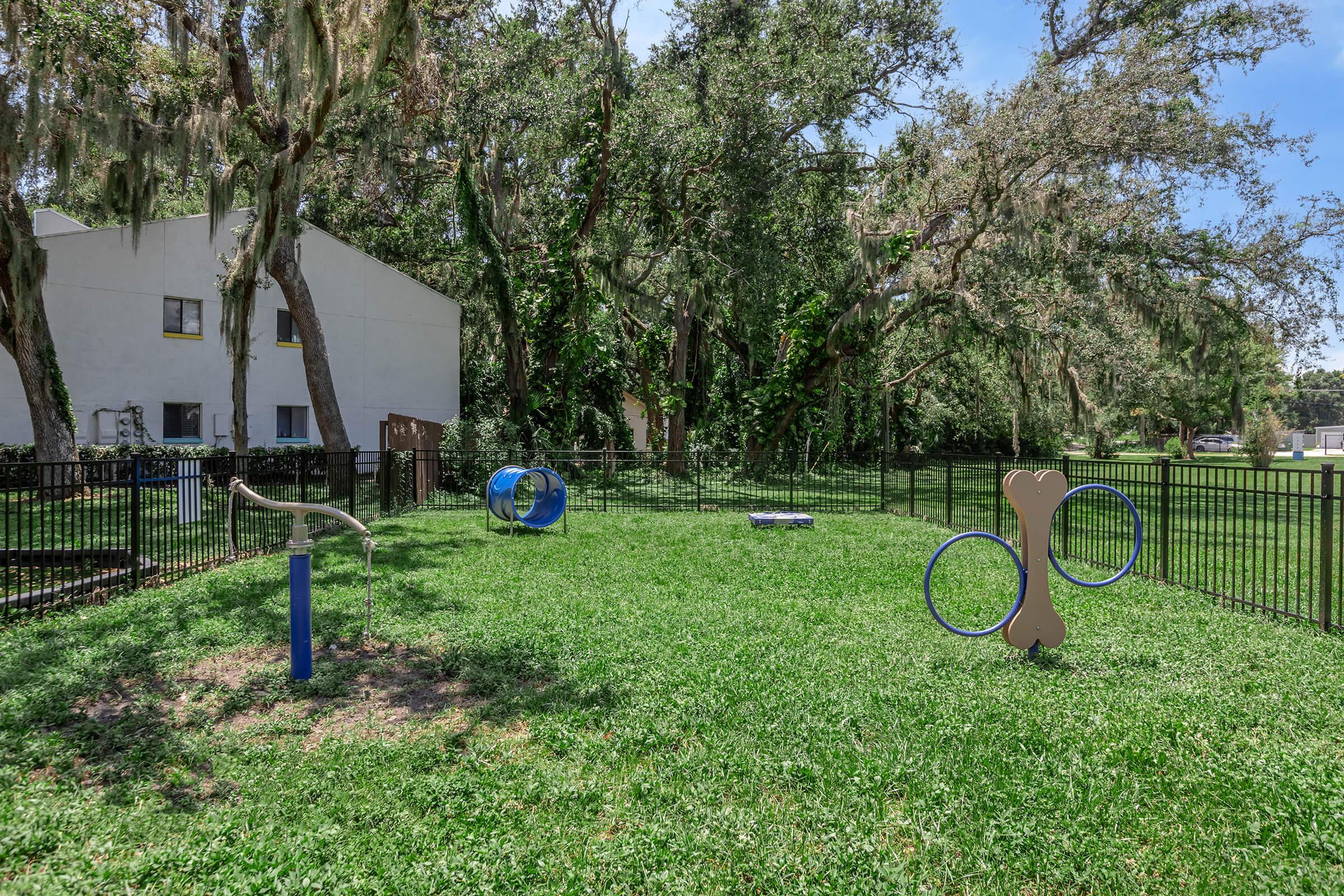 A grassy area with a fenced dog park featuring various agility equipment, including a tunnel, jumps, and a large bone-shaped structure, surrounded by trees and a residential building in the background.