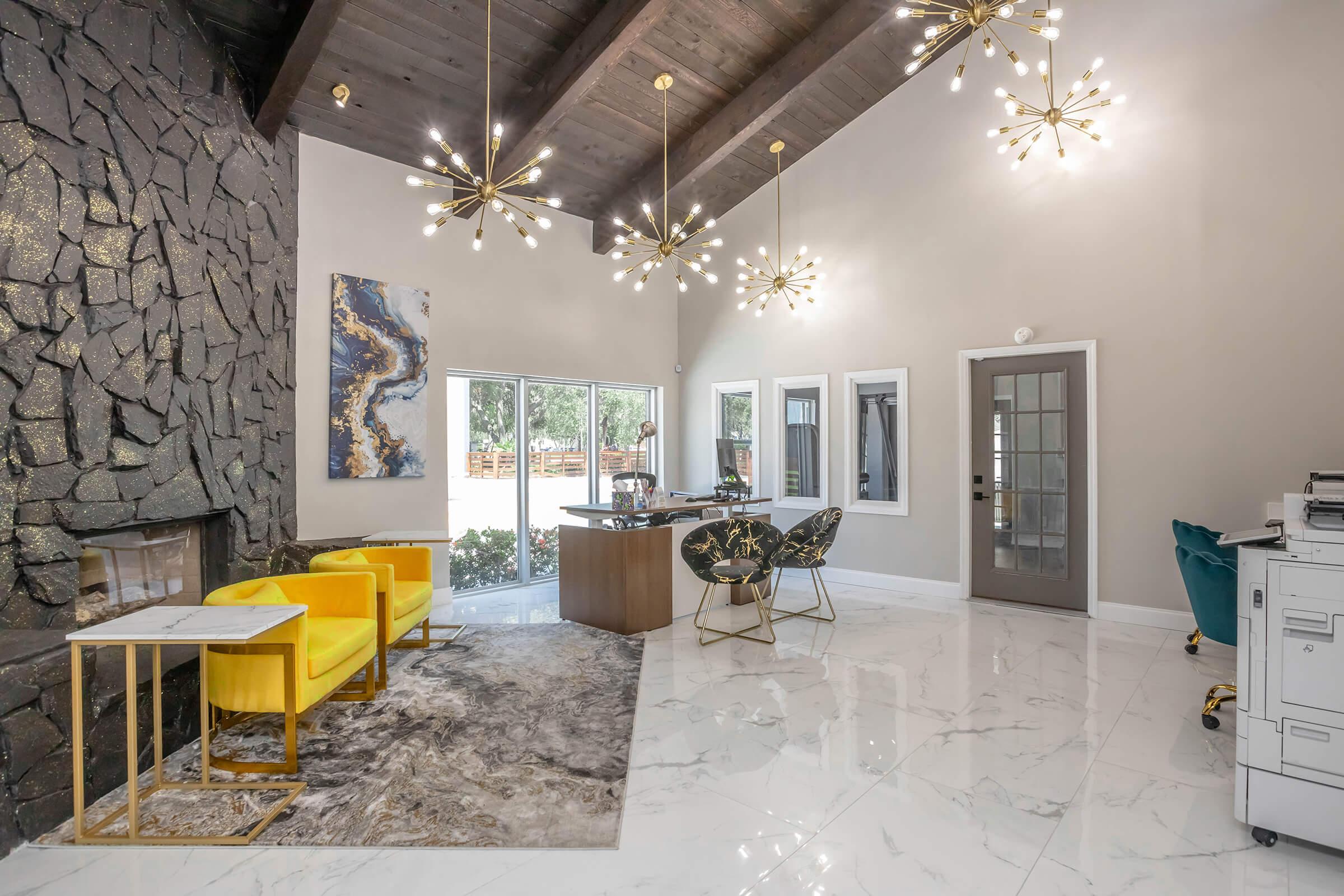 A modern interior space featuring a large stone wall with a fireplace, high ceilings with pendant lighting, yellow armchairs, and a stylish table. Large windows provide ample natural light, and a sleek desk is positioned in the center. The floor is glossy and white, adding to the contemporary aesthetic.