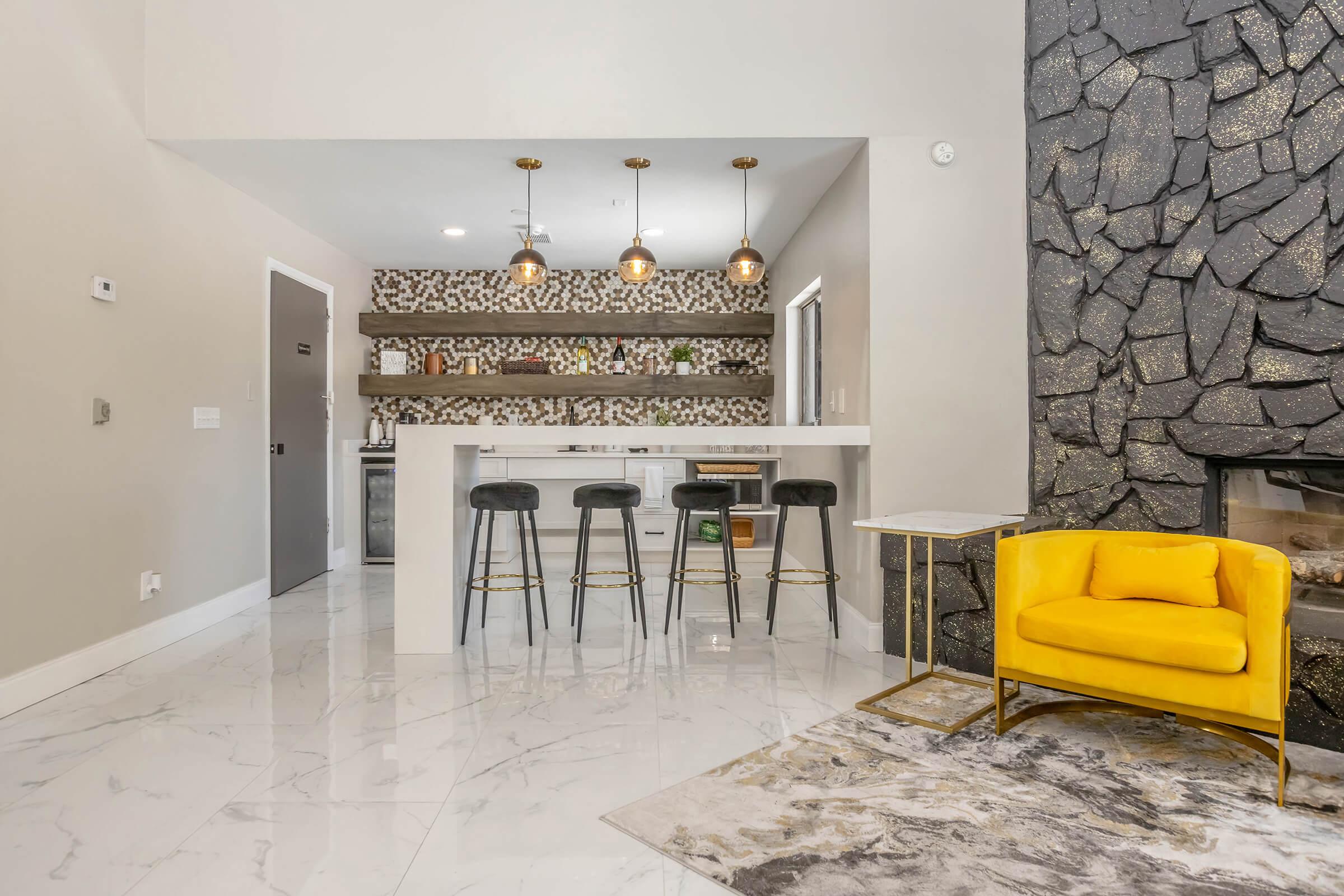 Modern interior featuring a sleek kitchen bar with three black stools, a decorative stone wall, and a bright yellow armchair. The space has marble flooring, warm lighting from pendant lamps, and a stylish rug. Shelves with green plants and decorative items add to the contemporary aesthetic.