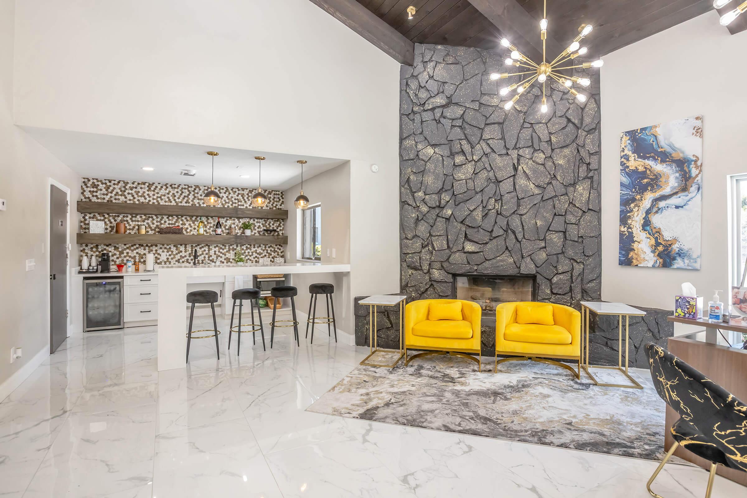 Modern living space featuring a cozy seating area with bright yellow chairs, a sleek marble floor, and a striking stone fireplace. The kitchen bar area, adorned with decorative tiles and stylish stools, complements the overall elegant design, while artistic decor adds a touch of sophistication.
