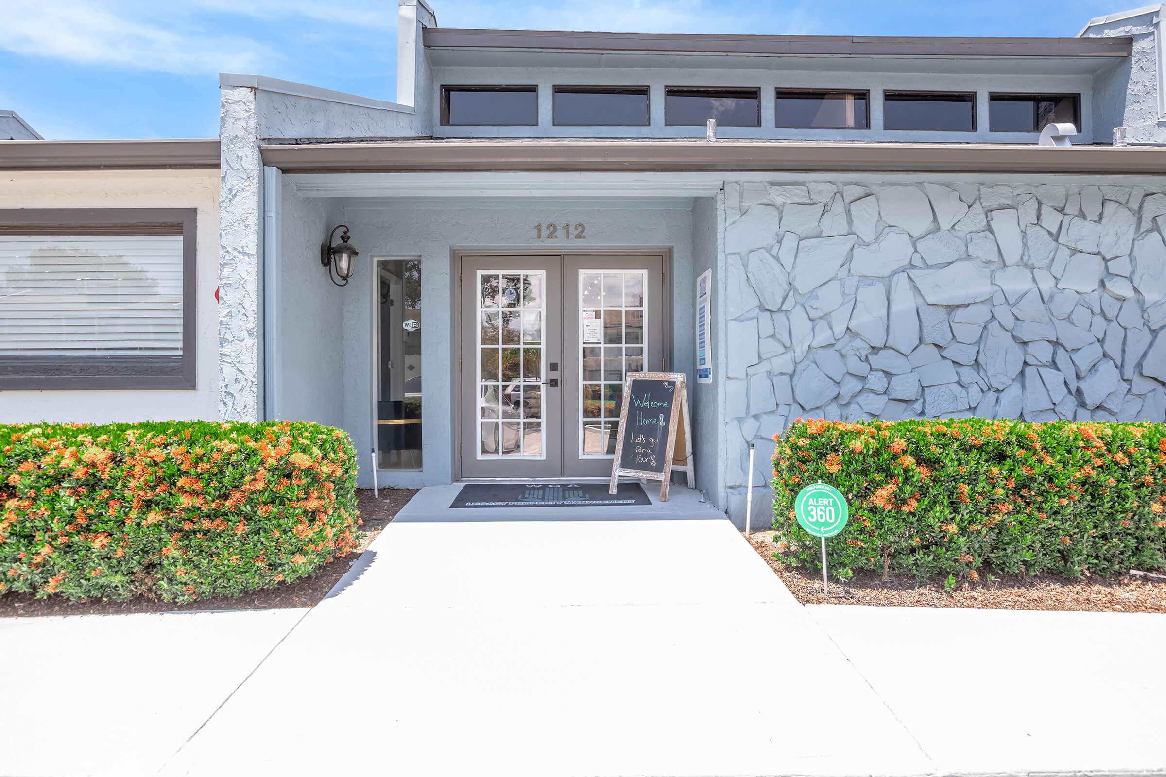Exterior view of a building with a stone facade, featuring a front entrance with glass doors. There is a small sign beside the door welcoming visitors, and neatly trimmed hedges with colorful flowers line the walkway. The address "1212" is visible above the door. Clear blue sky in the background.