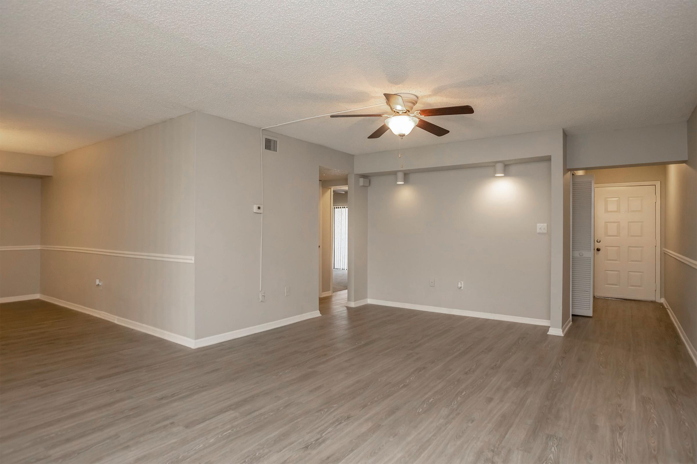 Spacious living area featuring light gray walls, textured ceiling, and a ceiling fan. The floor is finished with wood-like laminate. A doorway leads to another room, and there are wall-mounted light fixtures providing ambient lighting. The overall look is clean and modern, perfect for various interior design styles.