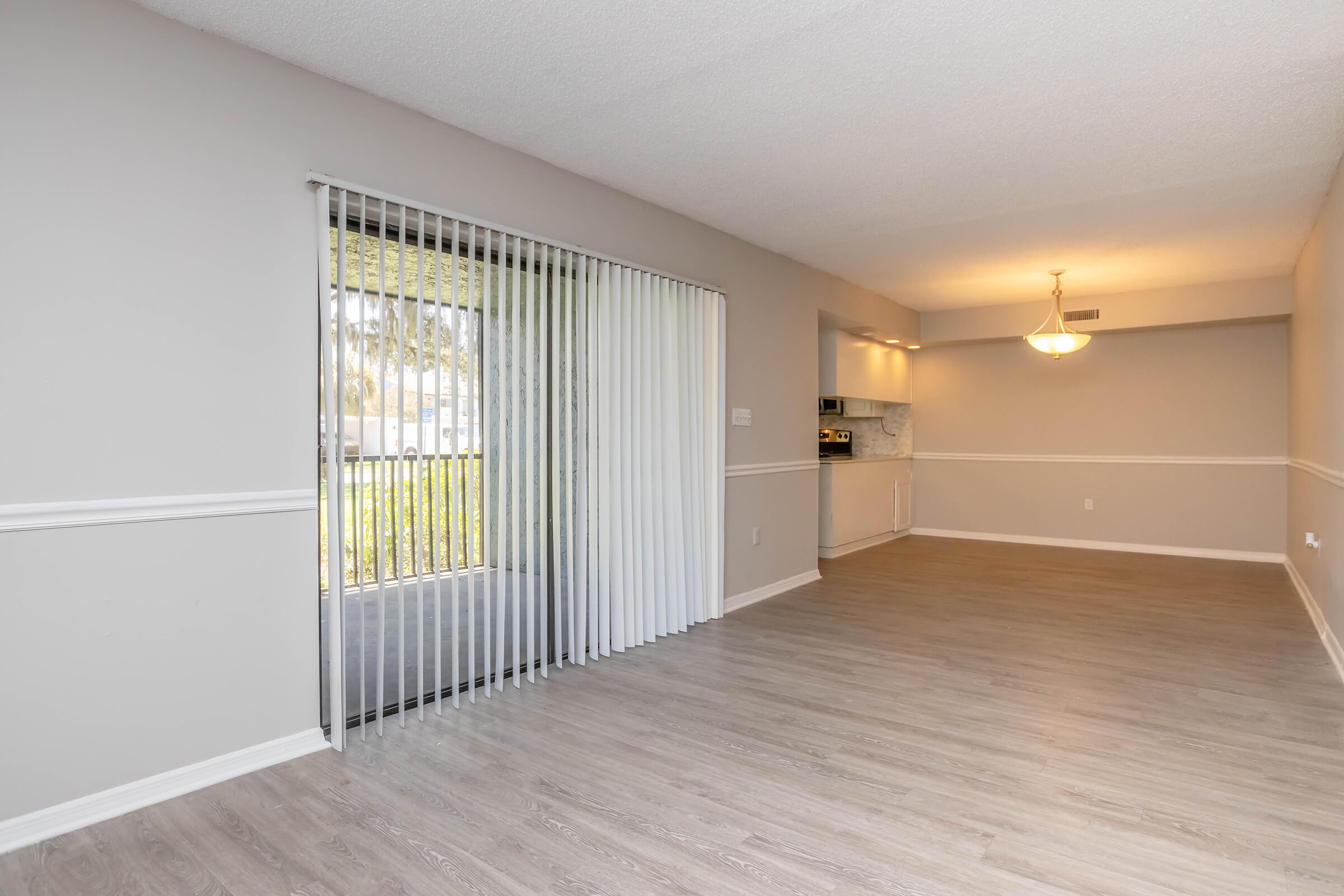 Spacious living area with a modern design, featuring light-colored walls and laminate flooring. A large window with vertical blinds allows natural light, and an open doorway leads to a kitchen area with a dining space illuminated by a pendant light.