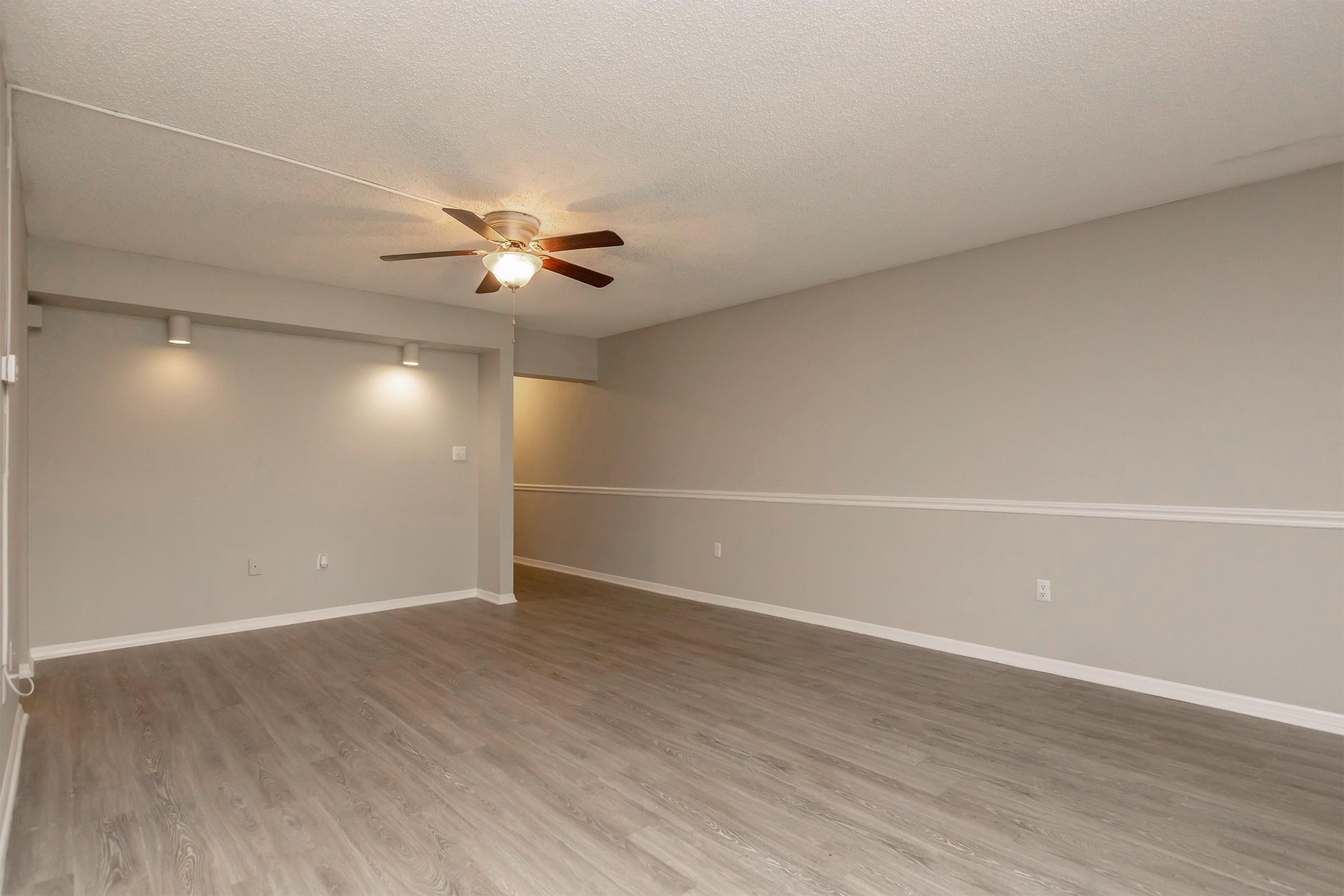 A spacious room with light gray walls and wood-like flooring. There is a ceiling fan in the center, and two wall sconces provide additional lighting. The room is empty, creating a bright and airy atmosphere, suitable for various interior design options.