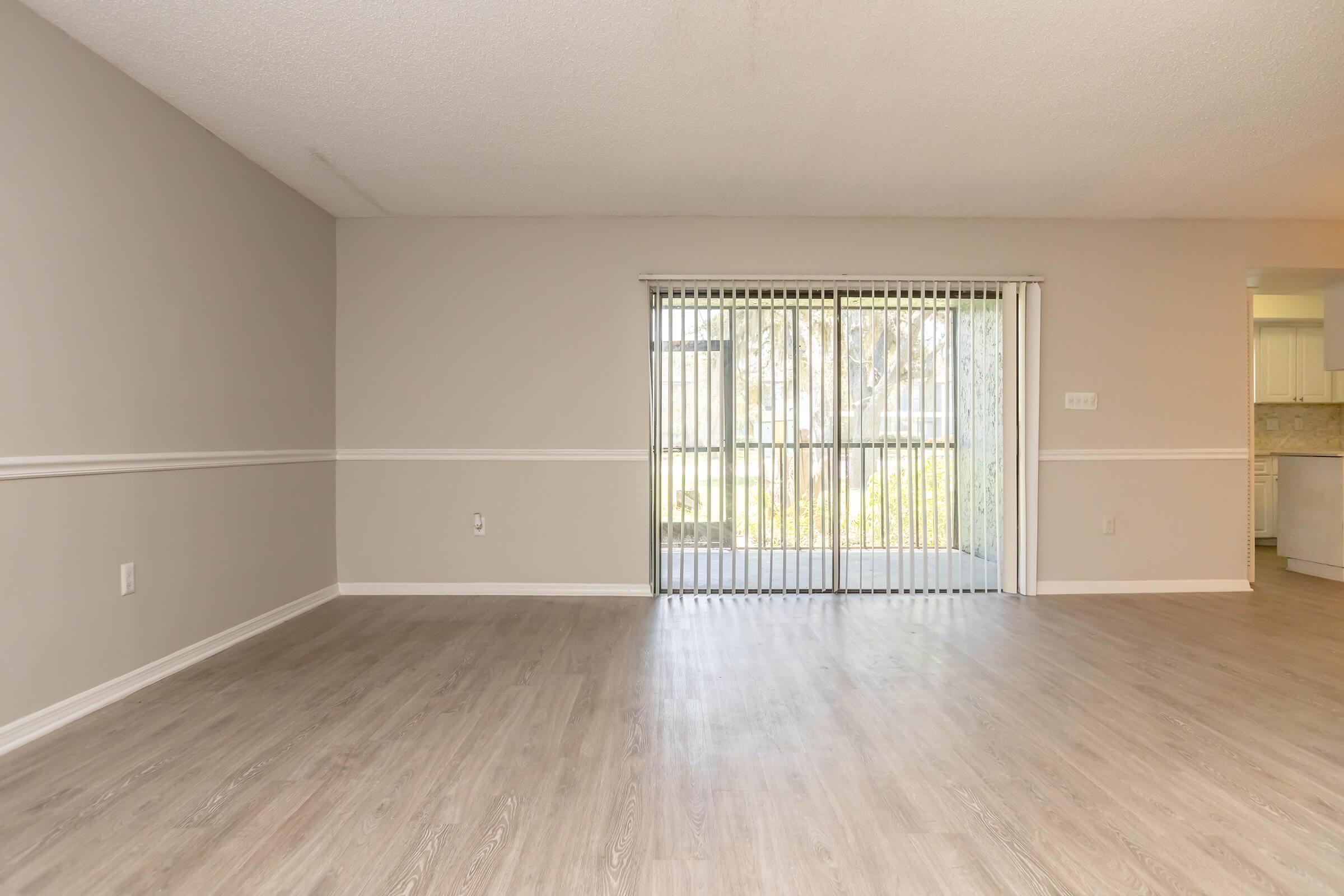 Spacious room with light-colored walls and wooden flooring, featuring a sliding glass door that leads to an outdoor area. The room has a decorative white chair rail and ample natural light from the outside. No furniture is present, emphasizing the open space.