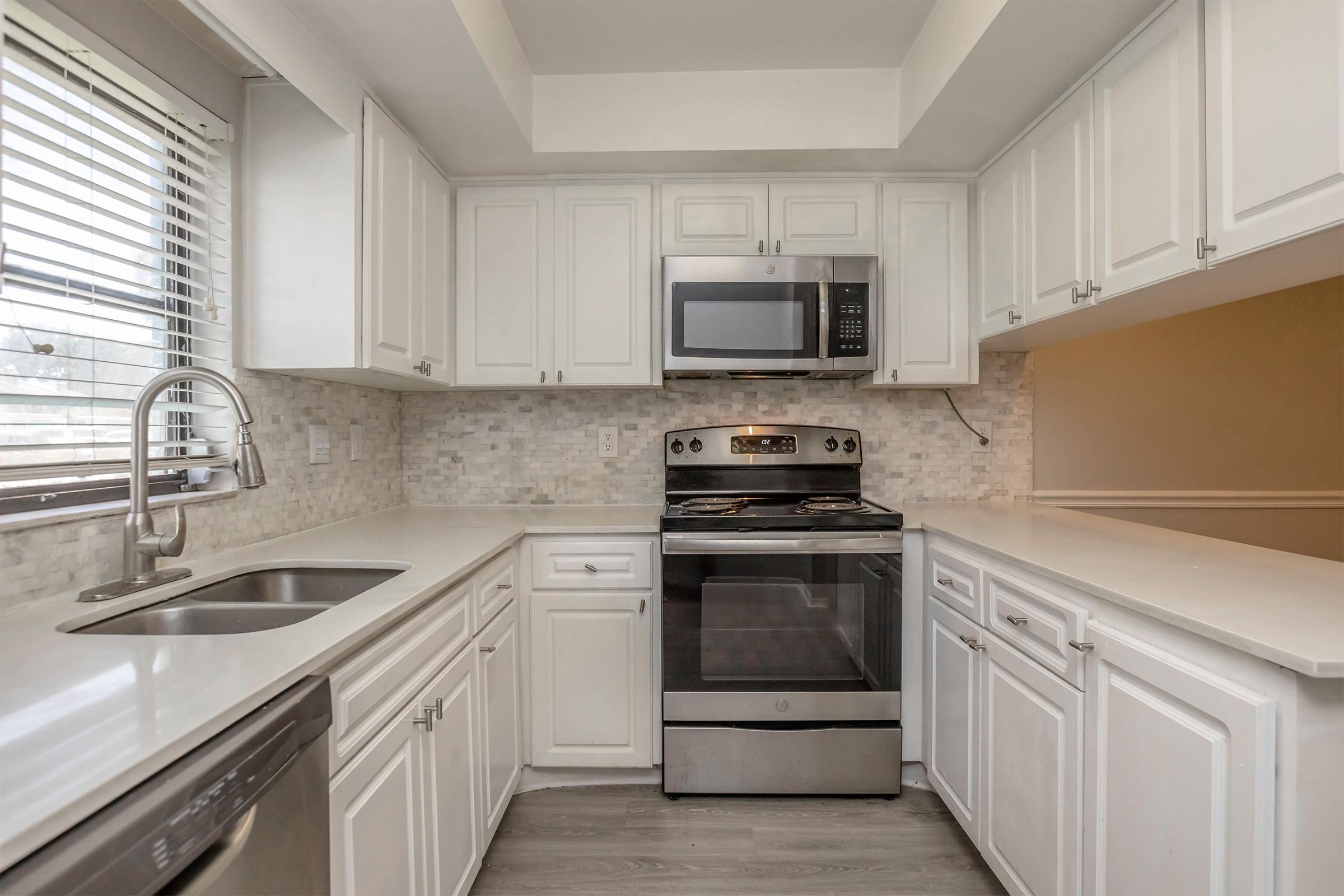 A modern kitchen featuring white cabinetry, stainless steel appliances including a microwave and oven, a silver sink, and light-colored countertops. There are windows with blinds allowing natural light, and a mosaic tile backsplash adds a stylish touch. The floor is a light wood laminate.