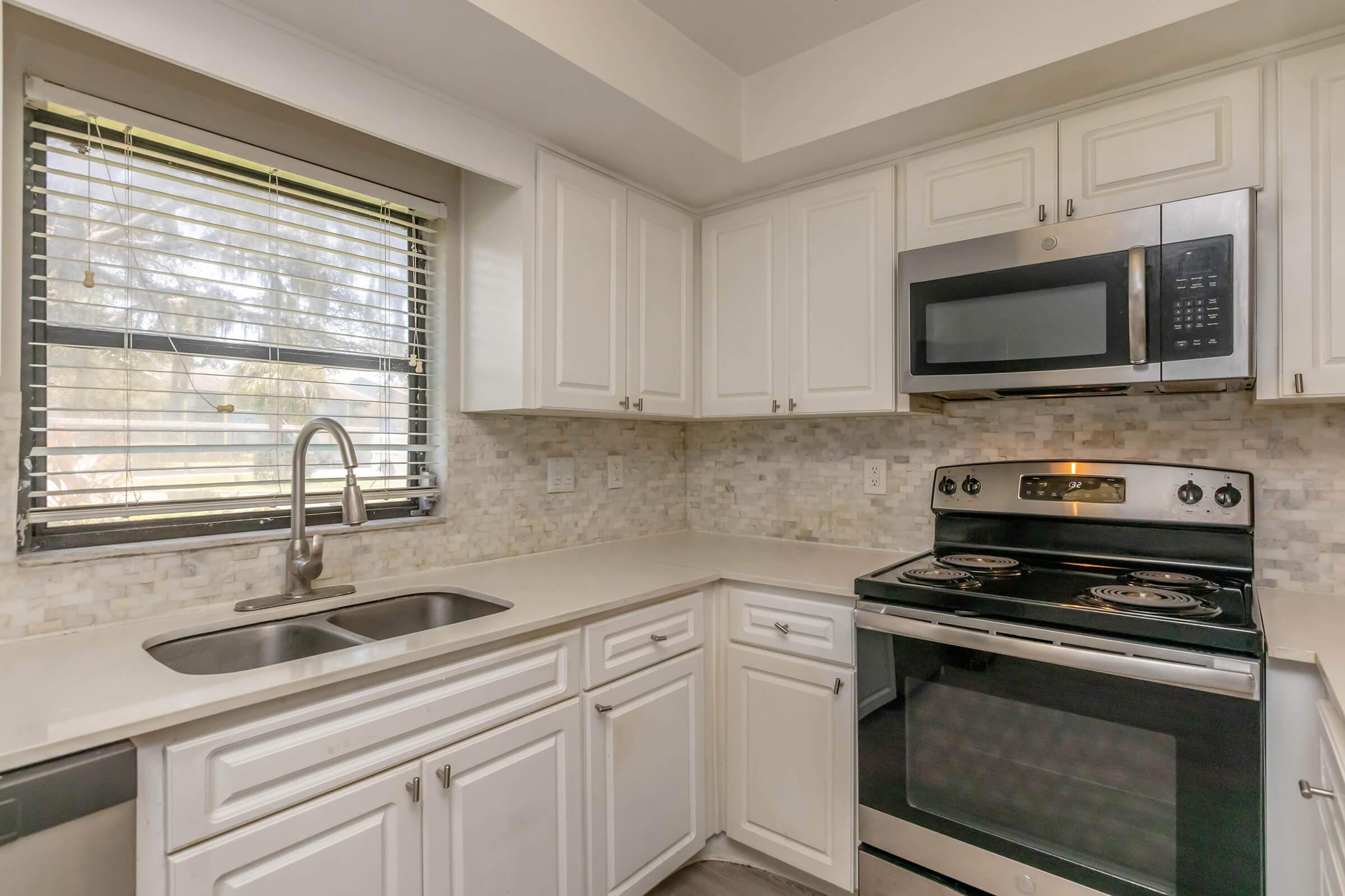Bright kitchen featuring white cabinets, a stainless steel microwave above an electric stove, and a double sink. The countertop is light-colored, and a textured backsplash adds visual interest. A window with blinds allows natural light to enter the space. The overall design is modern and inviting.