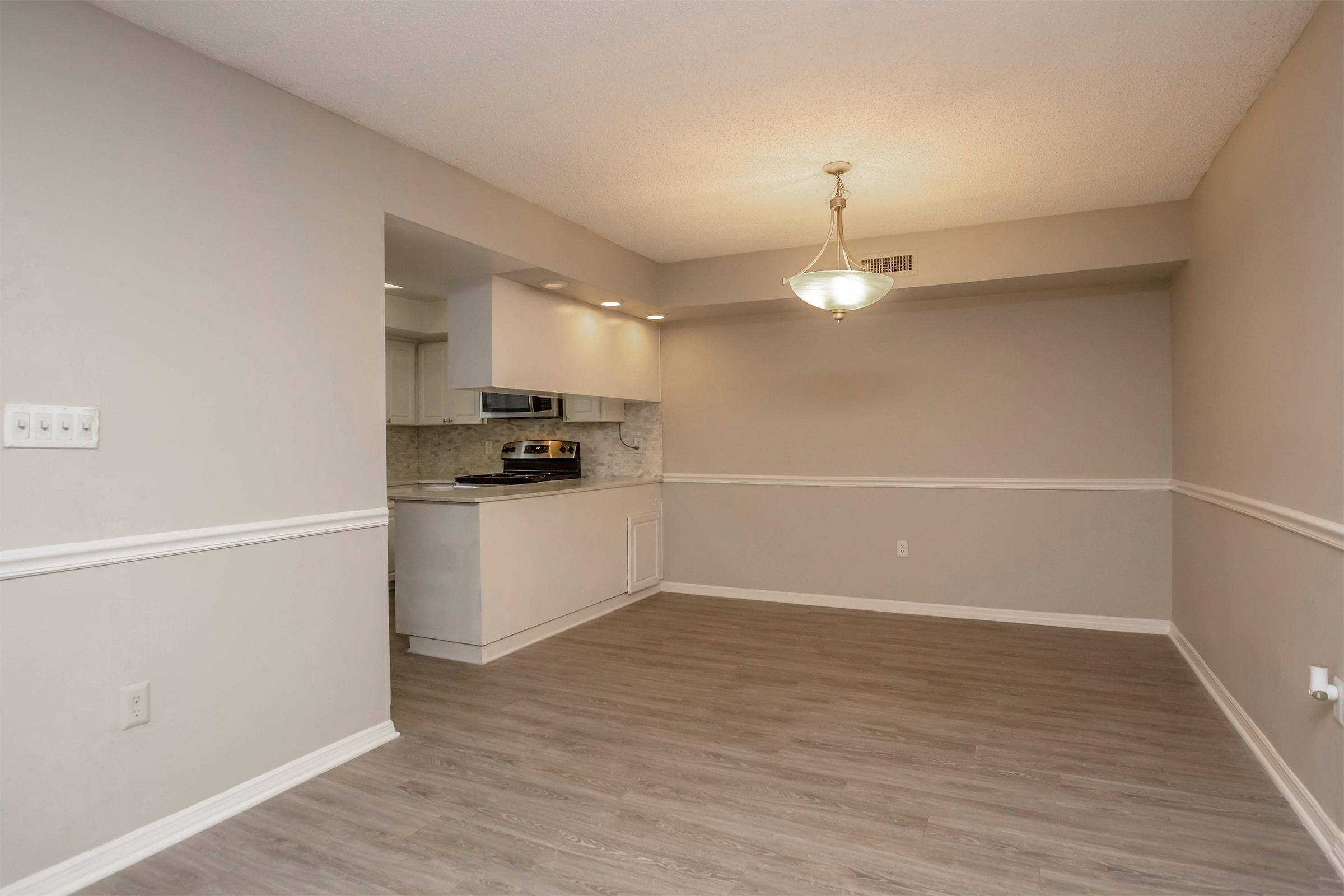 A spacious, modern living area featuring light gray walls and a subtle textured ceiling. The room includes a decorative chandelier and a half wall leading to a kitchen area with white cabinetry and a marble backsplash. The flooring is a light wood finish, enhancing the open and airy atmosphere.