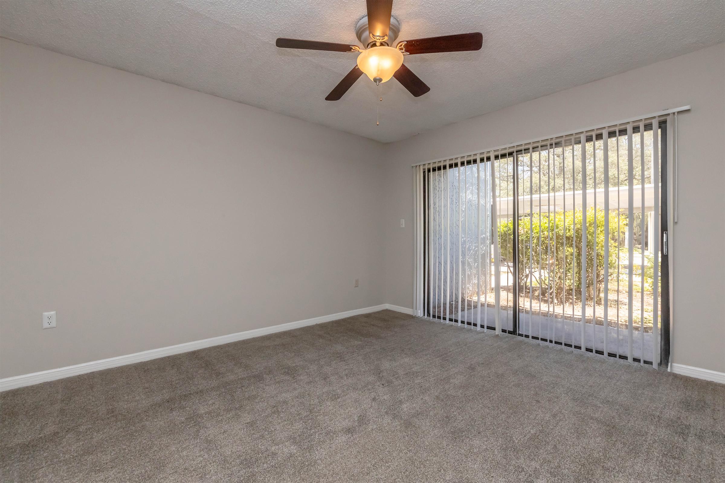 A spacious living room with light gray walls and a ceiling fan. The floor is covered in beige carpet, and there is a sliding glass door with vertical blinds leading to an outdoor area filled with greenery. The room has a bright and inviting atmosphere.