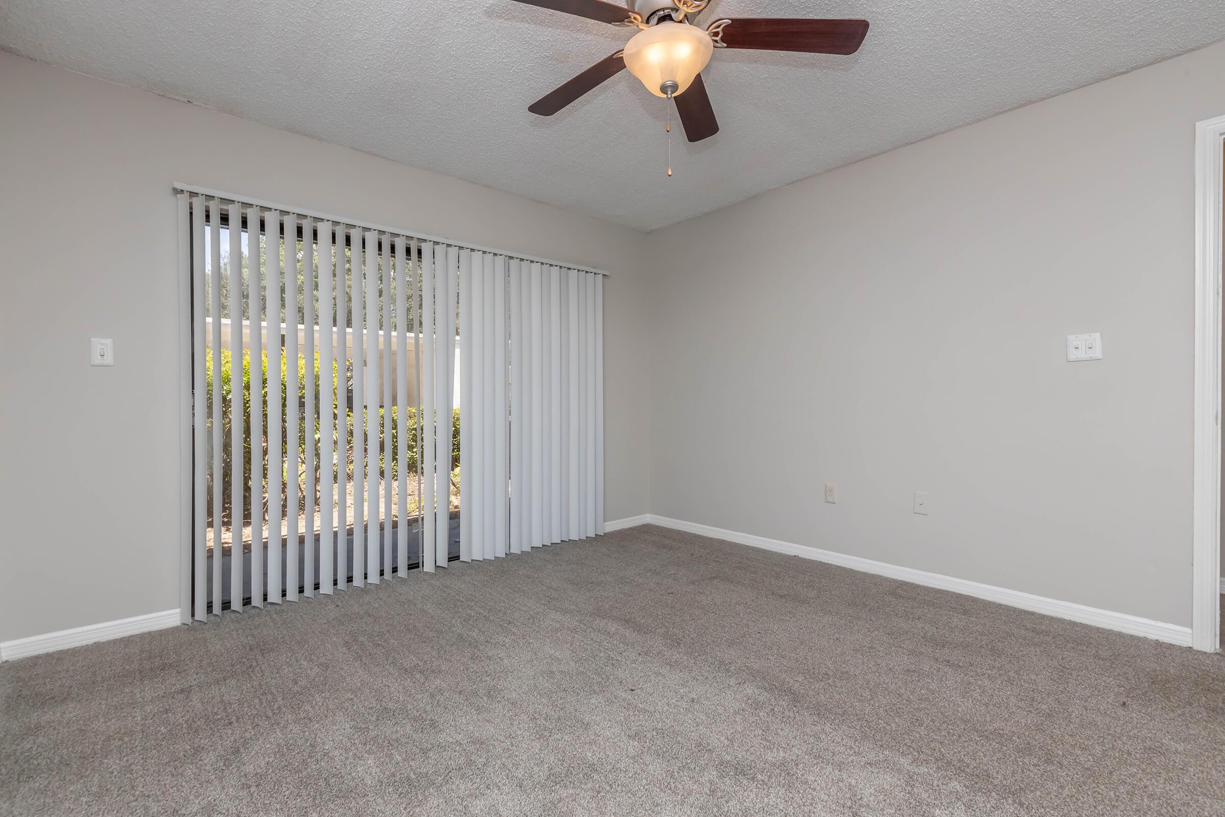 A spacious, empty room with light gray walls and a ceiling fan. Sunlight enters through vertical blinds, illuminating the plush, beige carpet. A door leads outside, and there are no furnishings, giving it an open, airy feel.