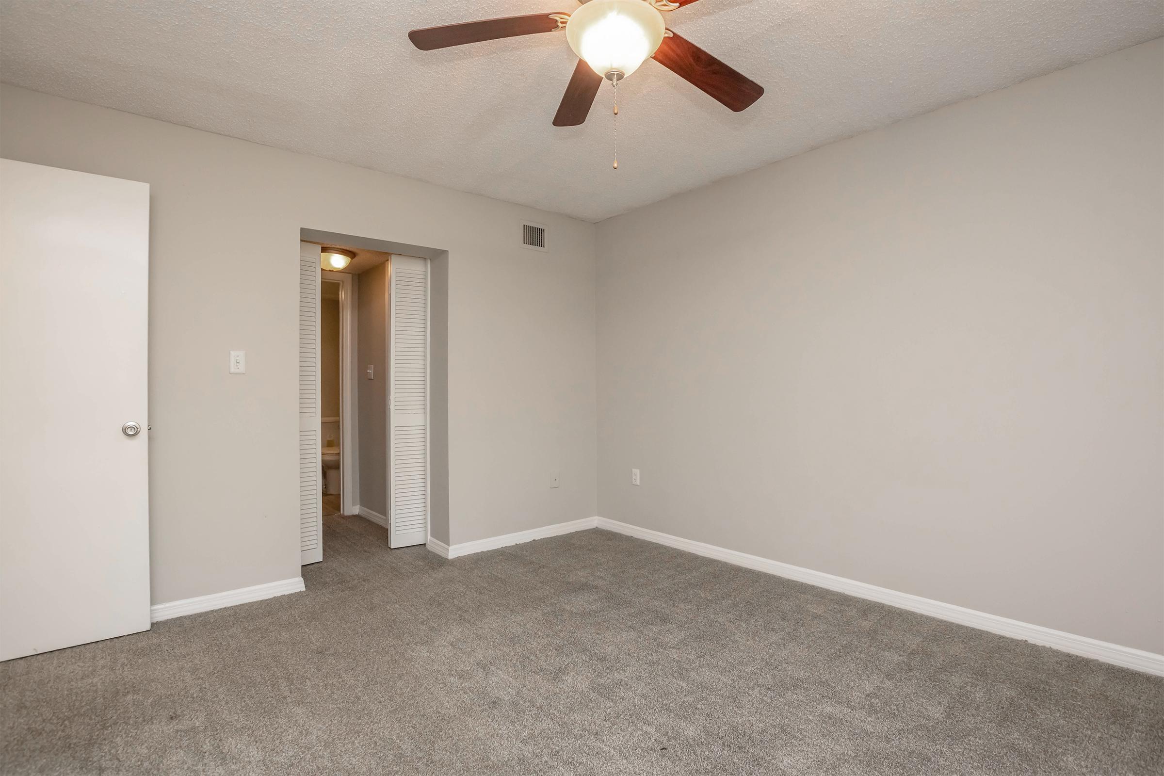 A well-lit, empty room with light gray walls and a ceiling fan. The floor is covered with carpet, and there is a door on the left leading to another room or hallway. The space is open and unadorned, suggesting it could be used for various purposes, such as a bedroom or office.
