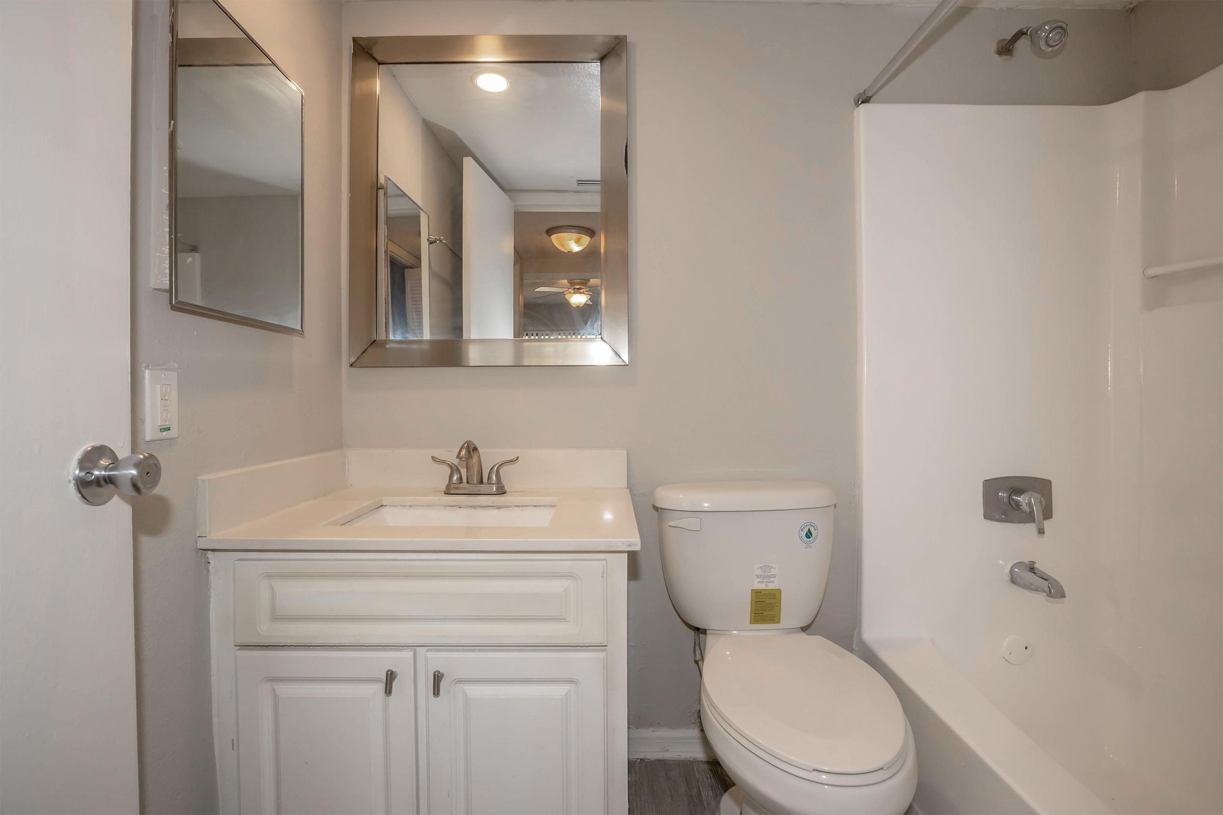 A clean and modern bathroom featuring a white sink with a mirror above, a toilet, and a bathtub with a shower. The walls are painted light gray, and there is a light fixture on the ceiling. The overall decor is simple and functional.
