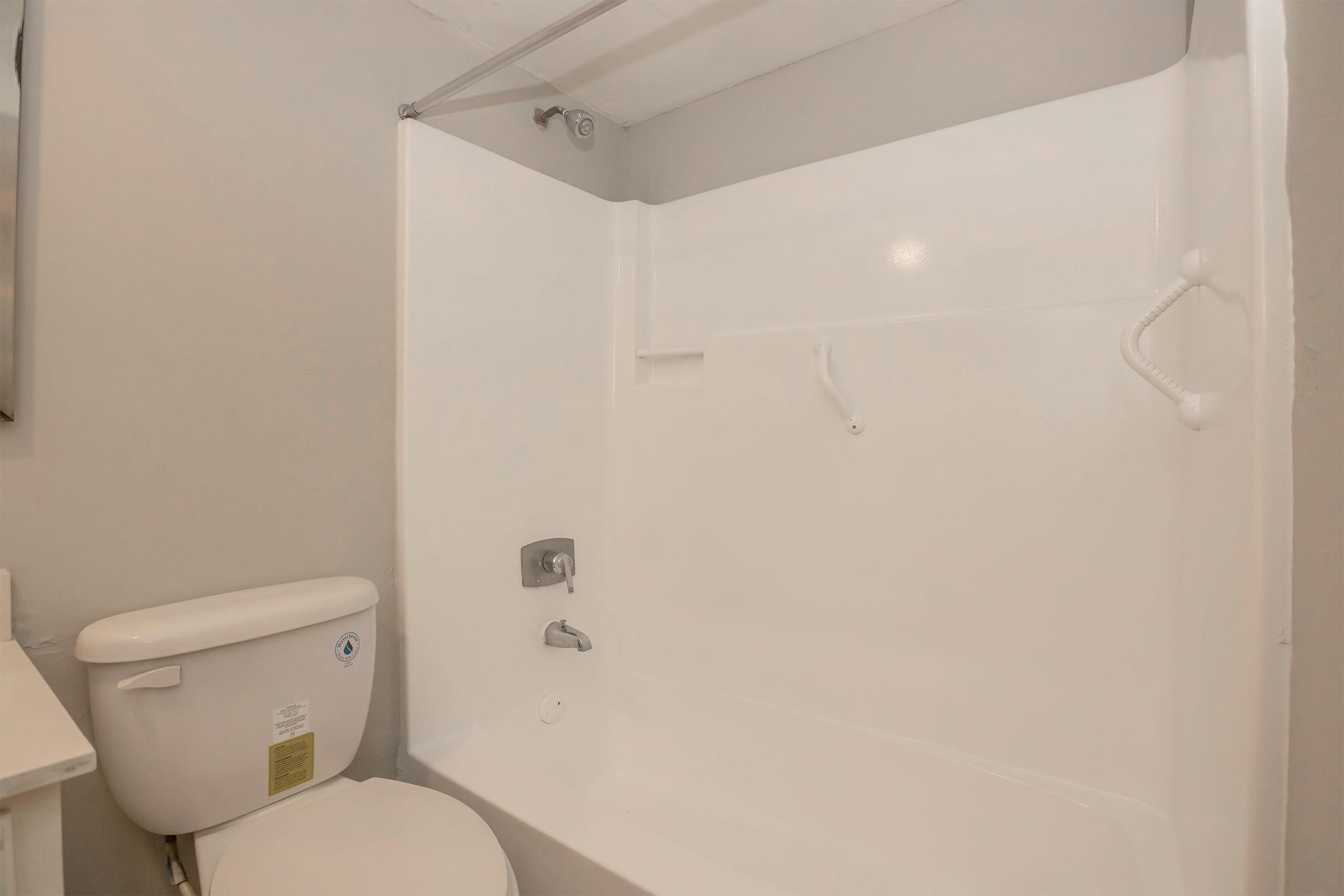 A clean bathroom featuring a white bathtub with a shower fixture, a toilet beside it, and light gray walls. The space is well-lit and shows a simple, modern design.