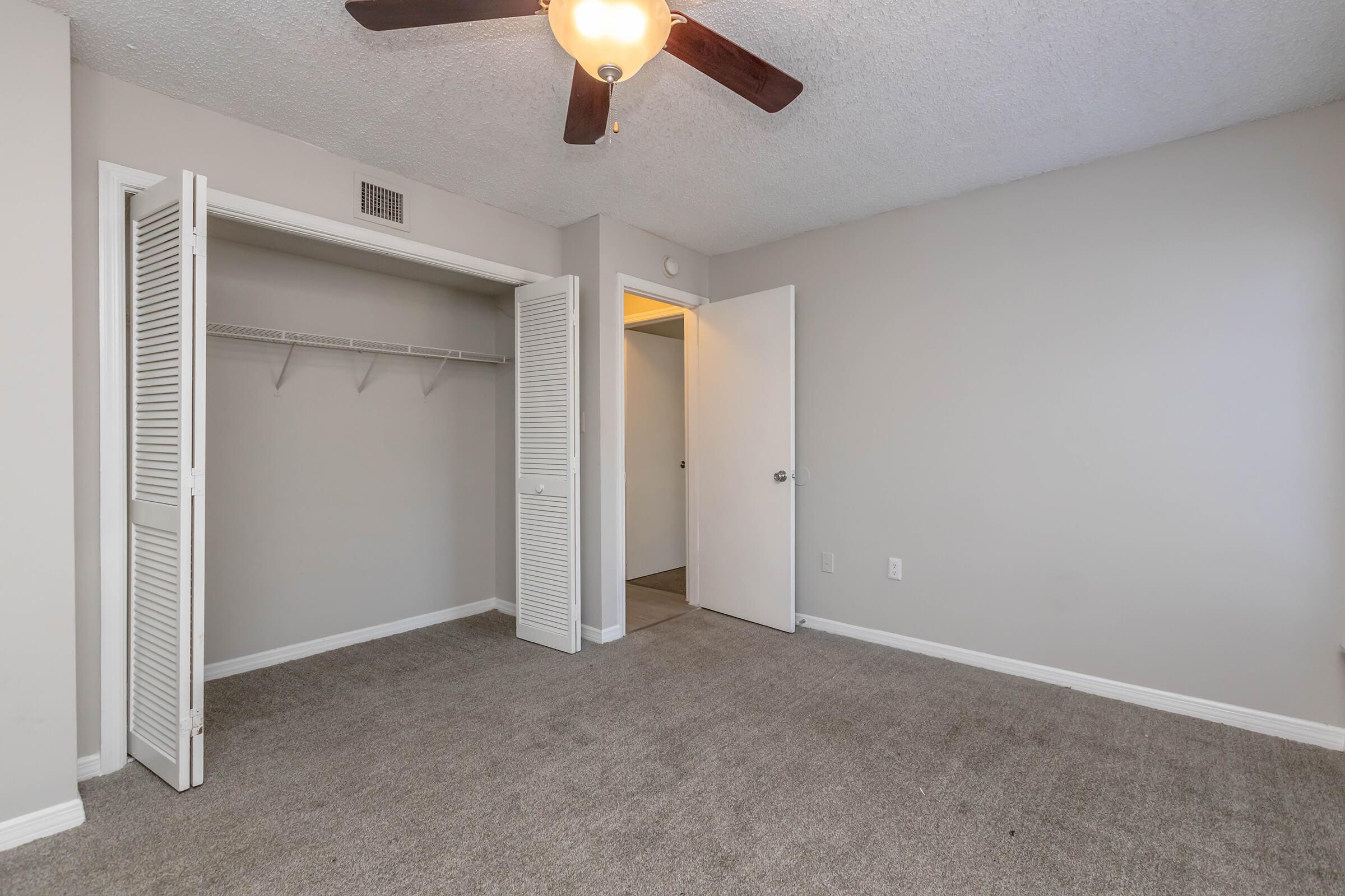 Empty room featuring light gray walls, a ceiling fan, and a carpeted floor. The room has a closet with sliding doors to the left and a doorway leading to another space. There's good natural light coming from the open door, creating a bright and airy atmosphere.