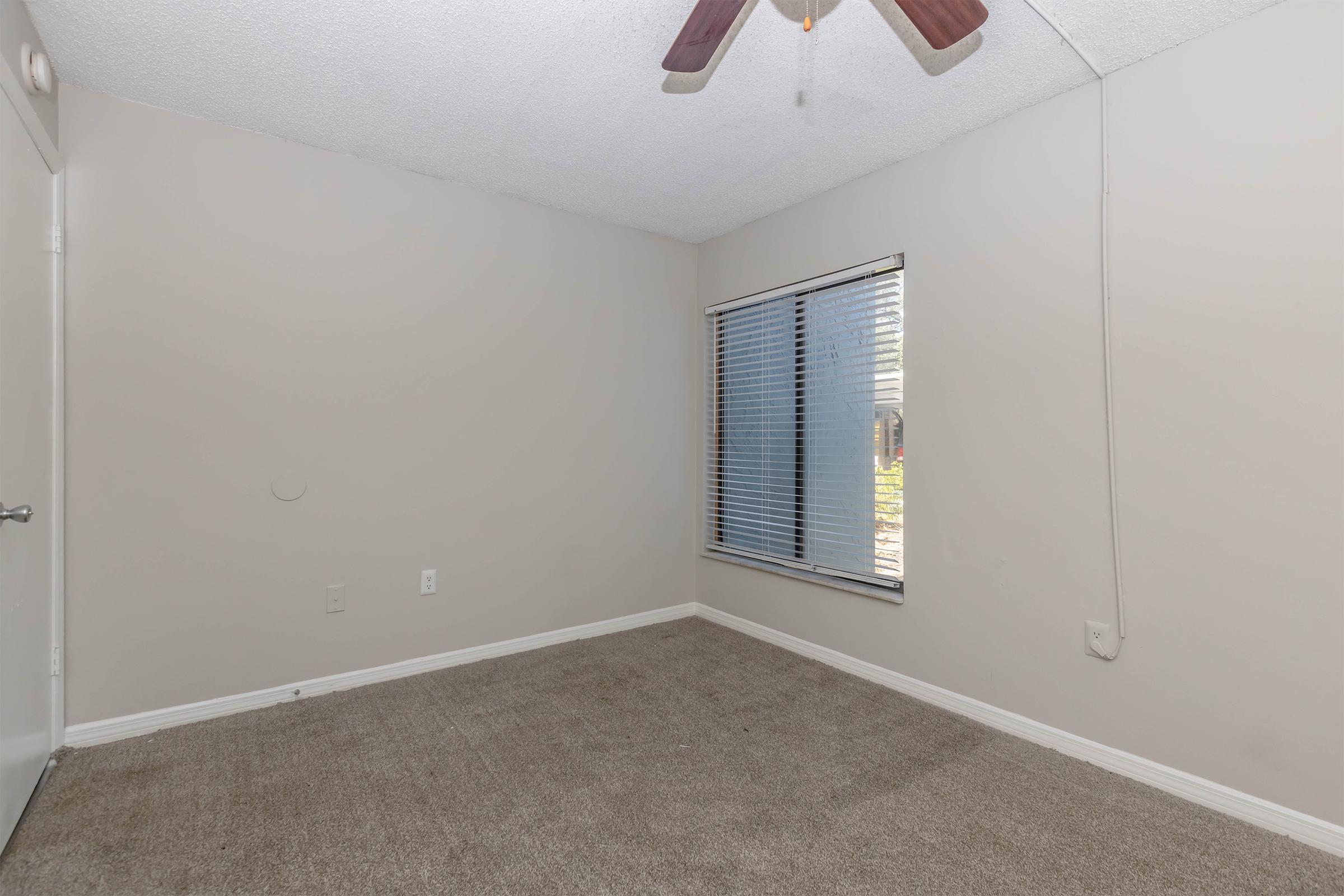 A small, empty room with light gray walls and a ceiling fan. There is a window with blinds allowing natural light to enter, and the floor is covered with carpet. The room appears to be unfurnished, creating a spacious and neutral environment.