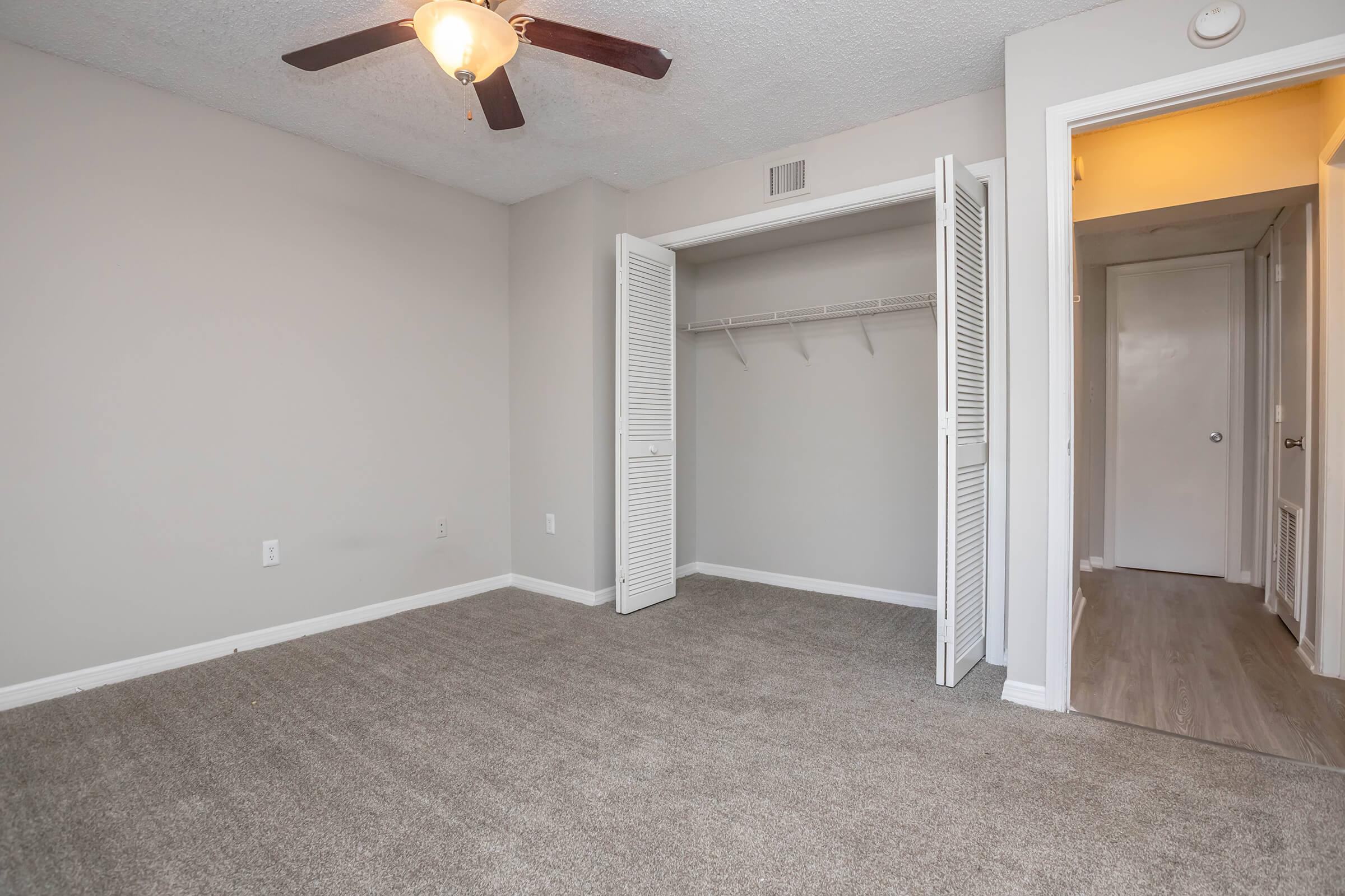 Empty room featuring beige walls and carpet, a ceiling fan, and a closet with doors opened to the left. A hallway can be seen to the right, leading to another area. Natural light is present in the space, creating a bright atmosphere.