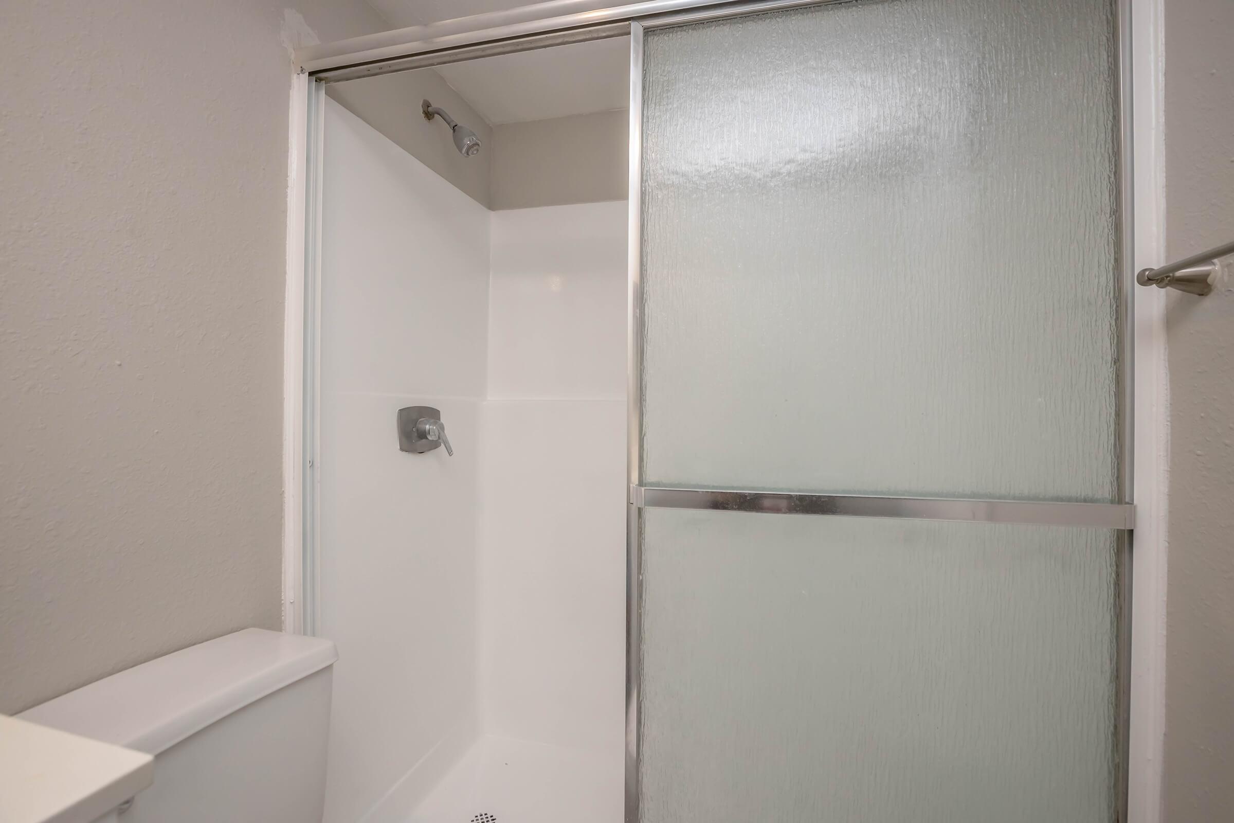 A clean, modern bathroom shower area with a glass door, featuring white walls and a silver showerhead. The adjoining toilet is visible, contributing to a spacious and bright appearance.