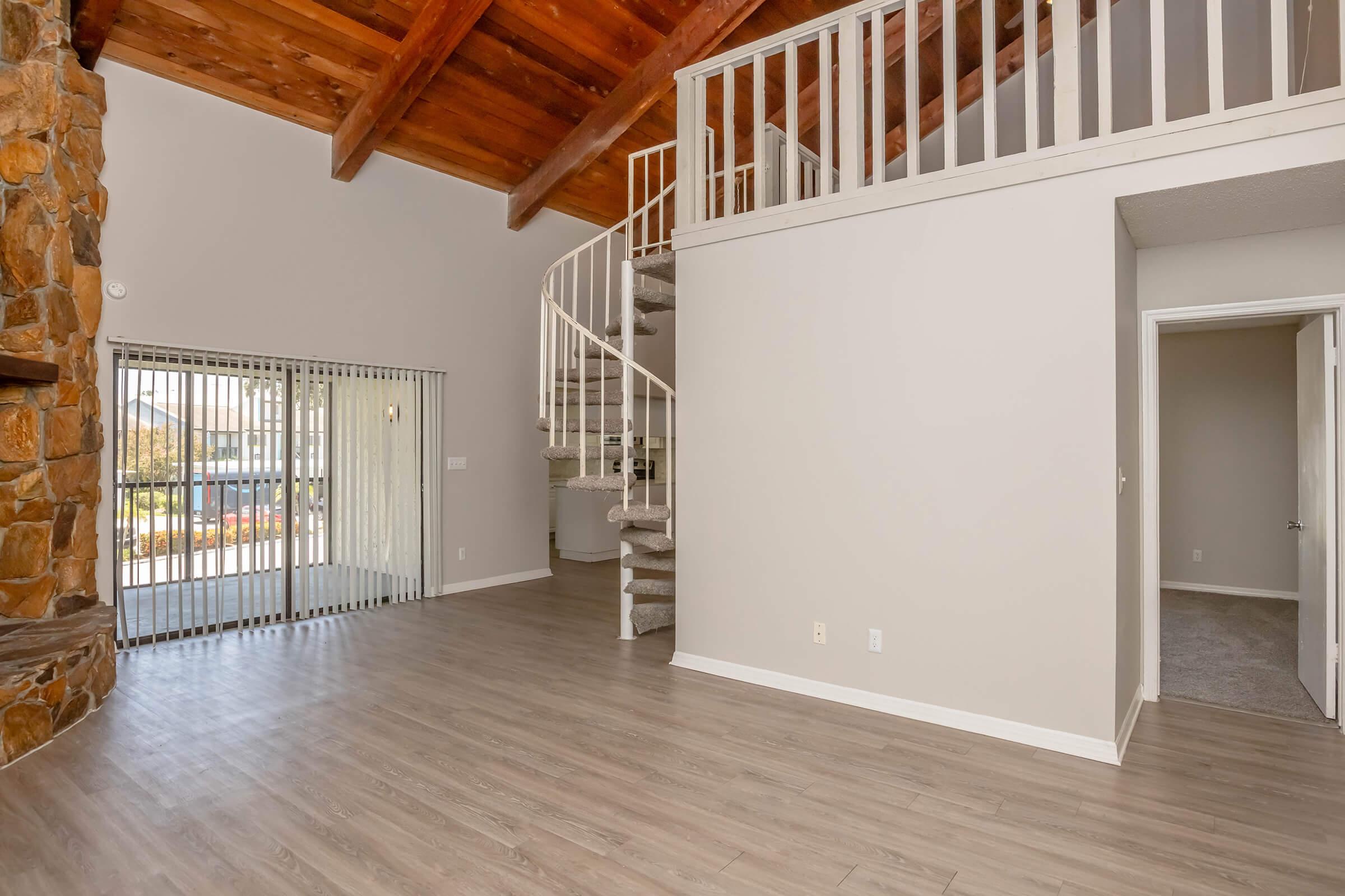 Interior of a spacious living area featuring a stone fireplace and a spiral staircase leading to a loft. The room has large windows with horizontal blinds and a neutral color scheme. The flooring is light wood, and there is an adjacent doorway leading to another room. The ceiling has exposed wooden beams.