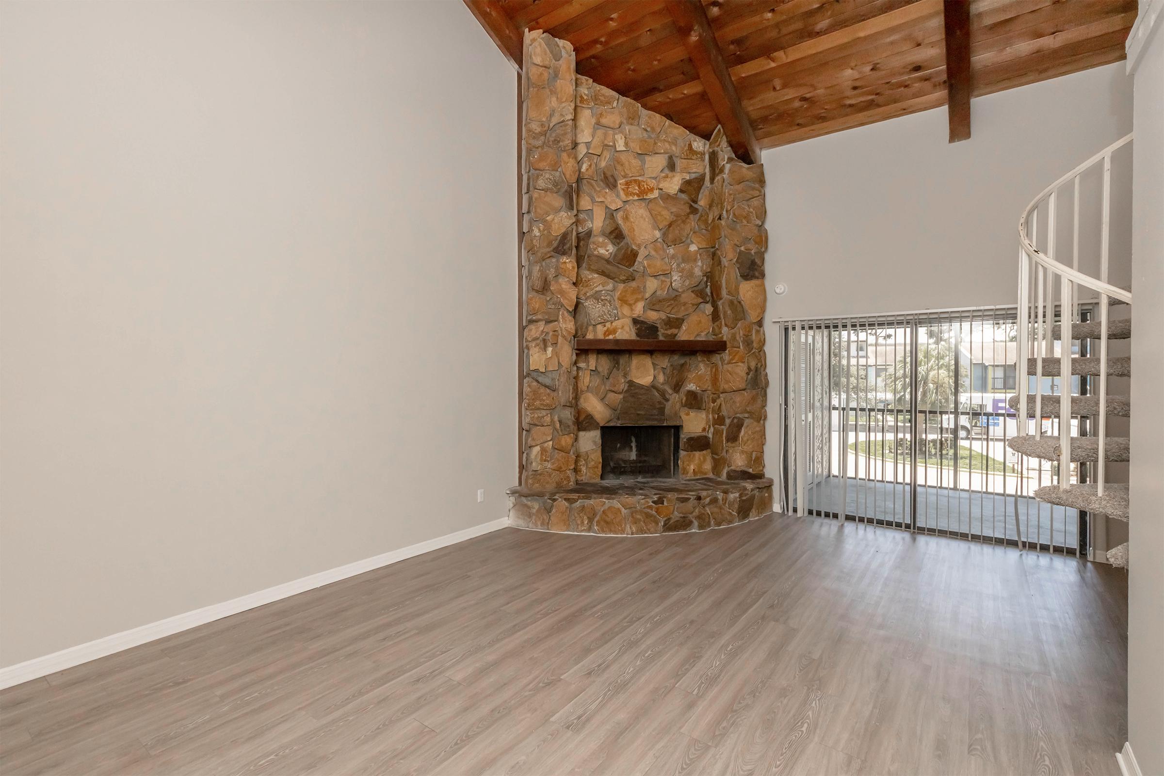 Spacious living room featuring a stone fireplace, high wooden beam ceiling, and large windows with views outside. The wood-style floor adds warmth to the space, while a spiral staircase on the right leads to an upper level. The walls are painted in a neutral color, enhancing the room's openness and brightness.