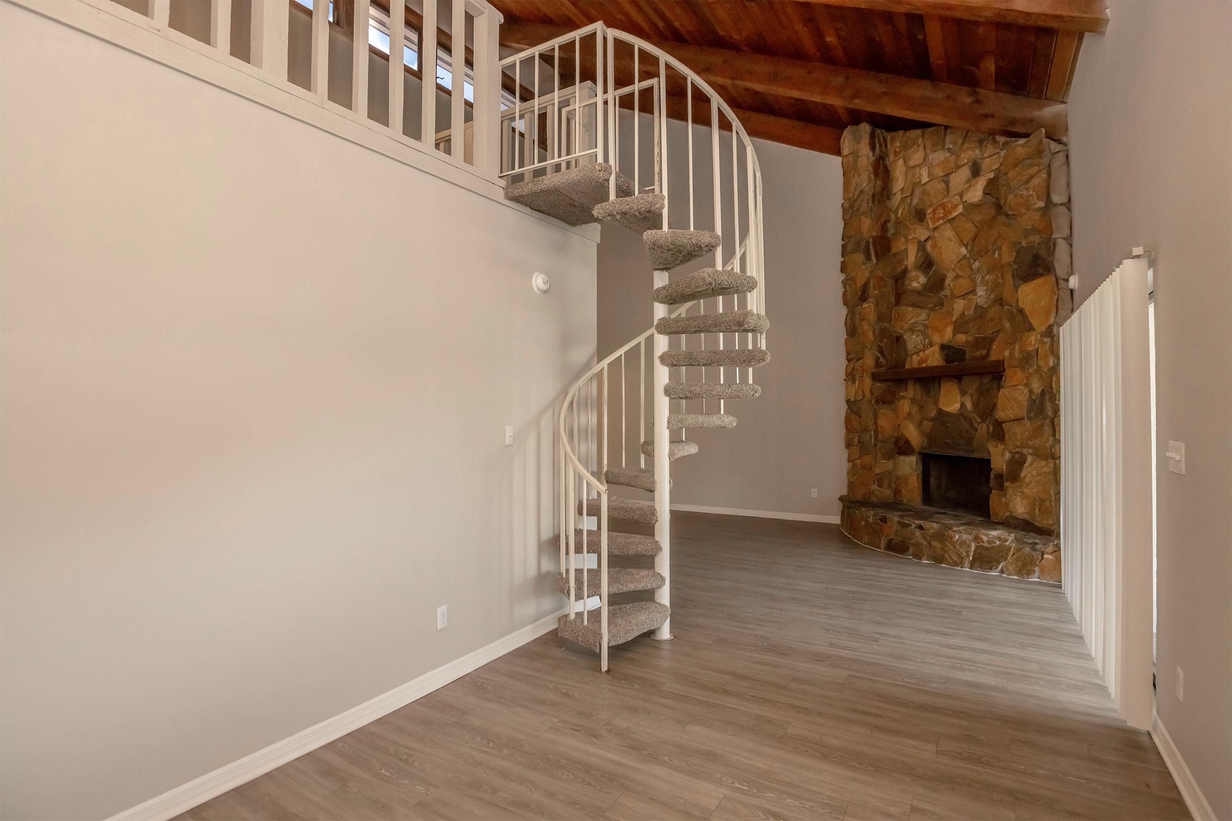 Interior view of a spacious room featuring a spiral staircase made of carpeted steps, a stone fireplace with a wooden mantel, and light-colored walls. The ceiling has wooden beams, and the flooring is a light wood laminate. Natural light enters through windows, enhancing the open atmosphere.