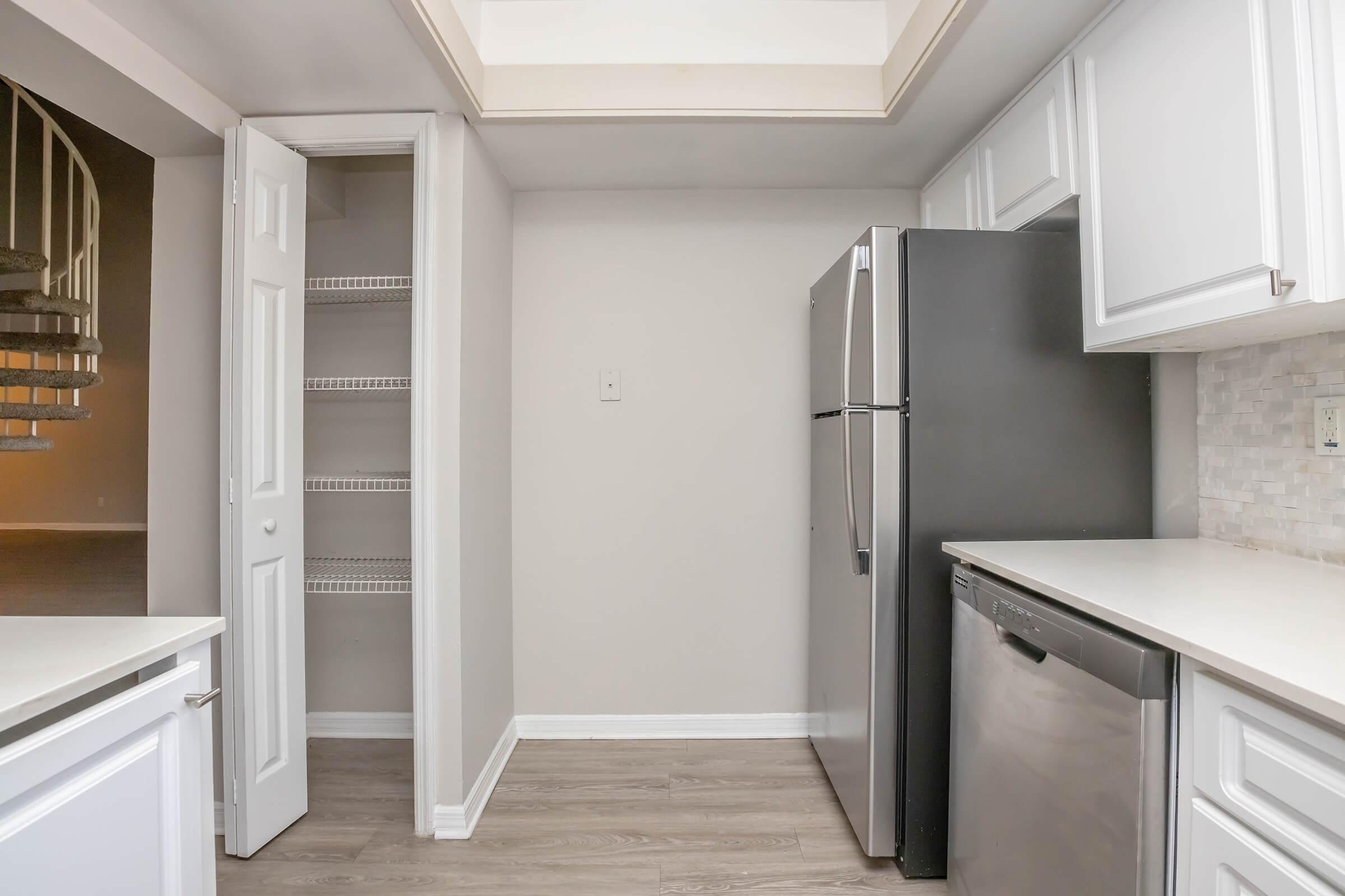 A modern kitchen featuring white cabinets, a stainless steel refrigerator, and a dishwasher. There is a small pantry with a white door and a ceiling with a skylight. The floor is a light wood finish, and the space is well-lit, creating a clean and inviting atmosphere.