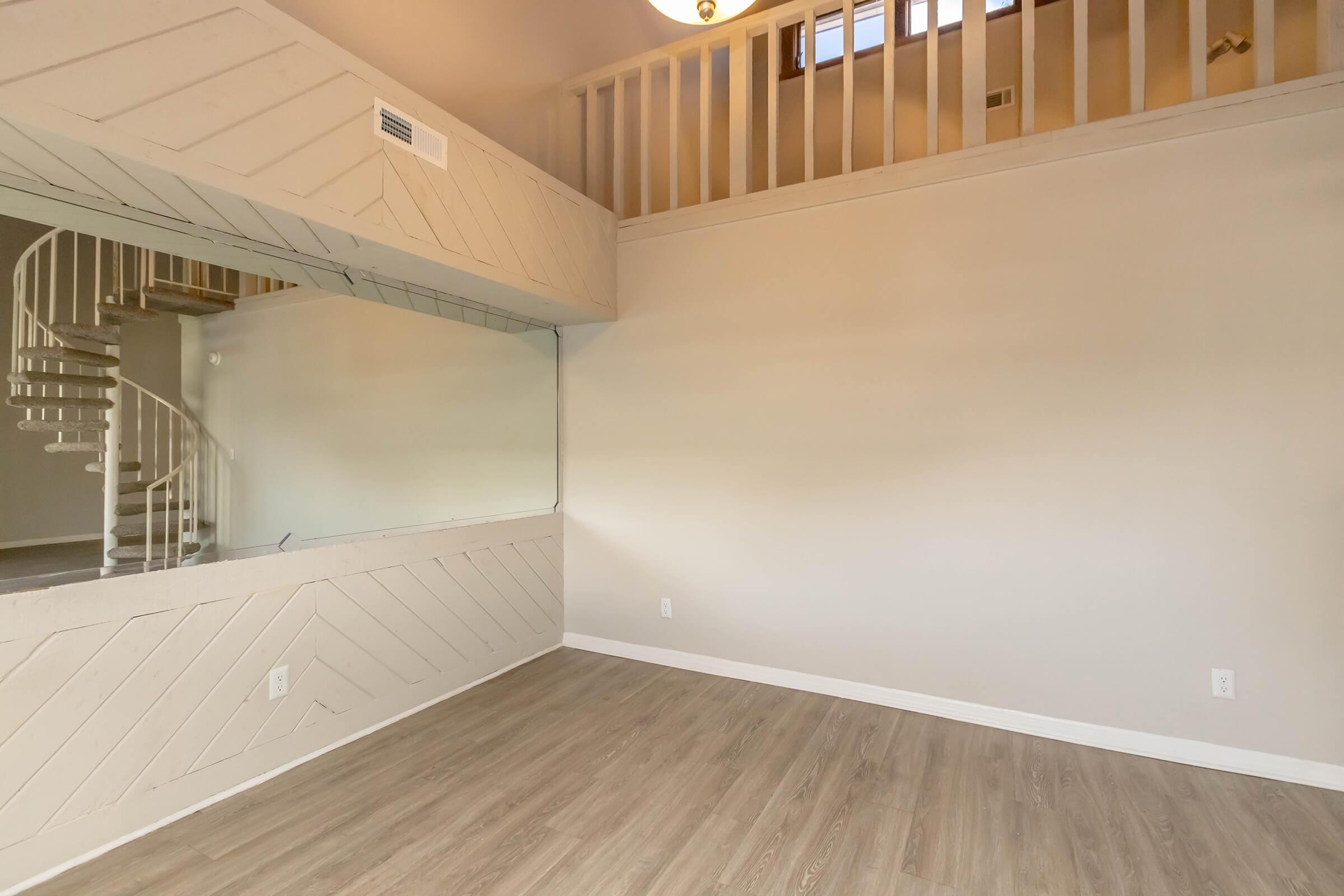 An empty interior space featuring light-colored walls and wooden flooring. There is a spiral staircase in the background leading to an upper level, and a large mirror on one wall reflecting the room. The space has a modern, open layout with plenty of natural light.