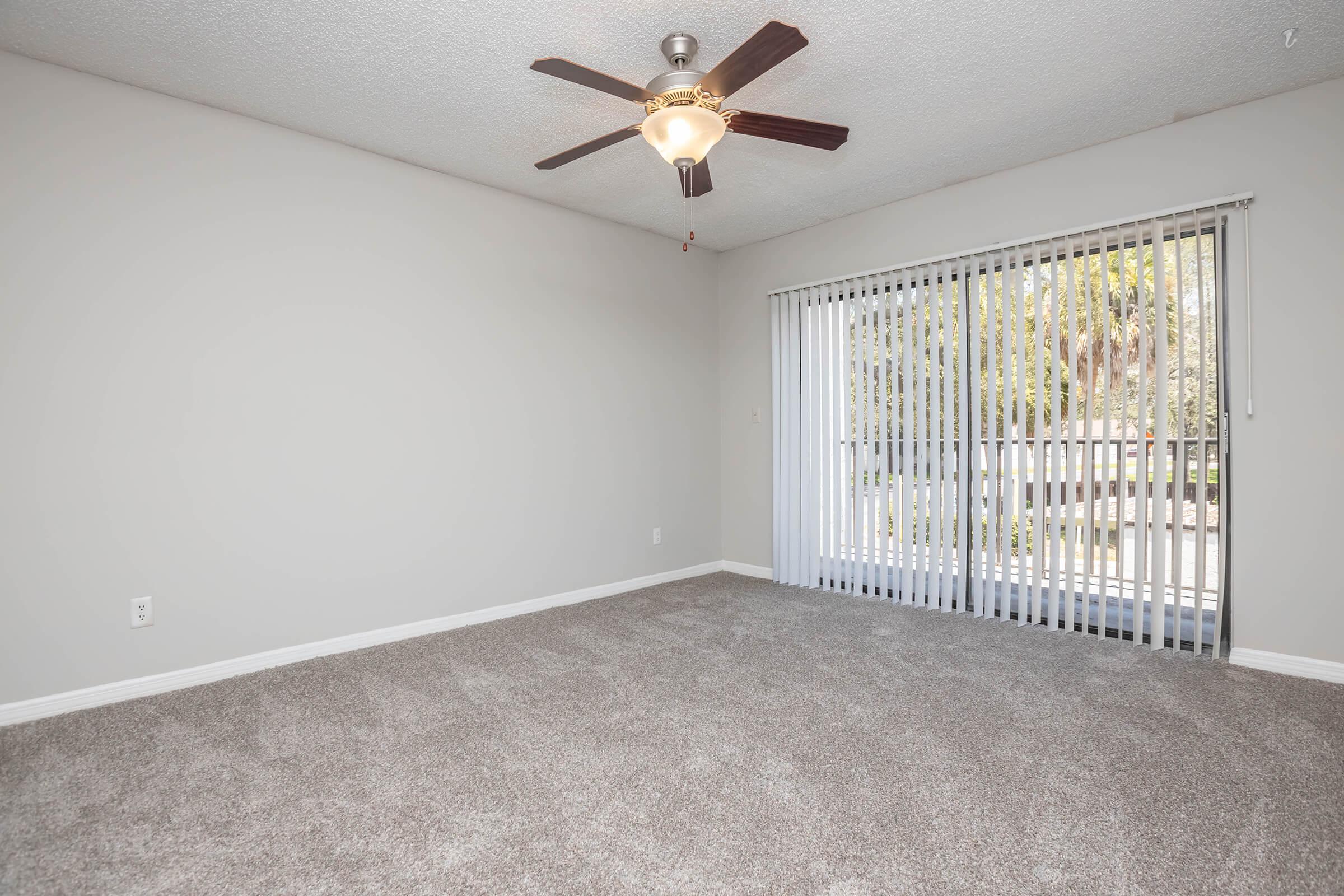 A spacious, empty room with light gray walls and soft carpet. It features a ceiling fan with wooden blades, and a large window with vertical blinds allowing natural light to enter. The room has a clean, modern look, perfect for personalization and decoration.