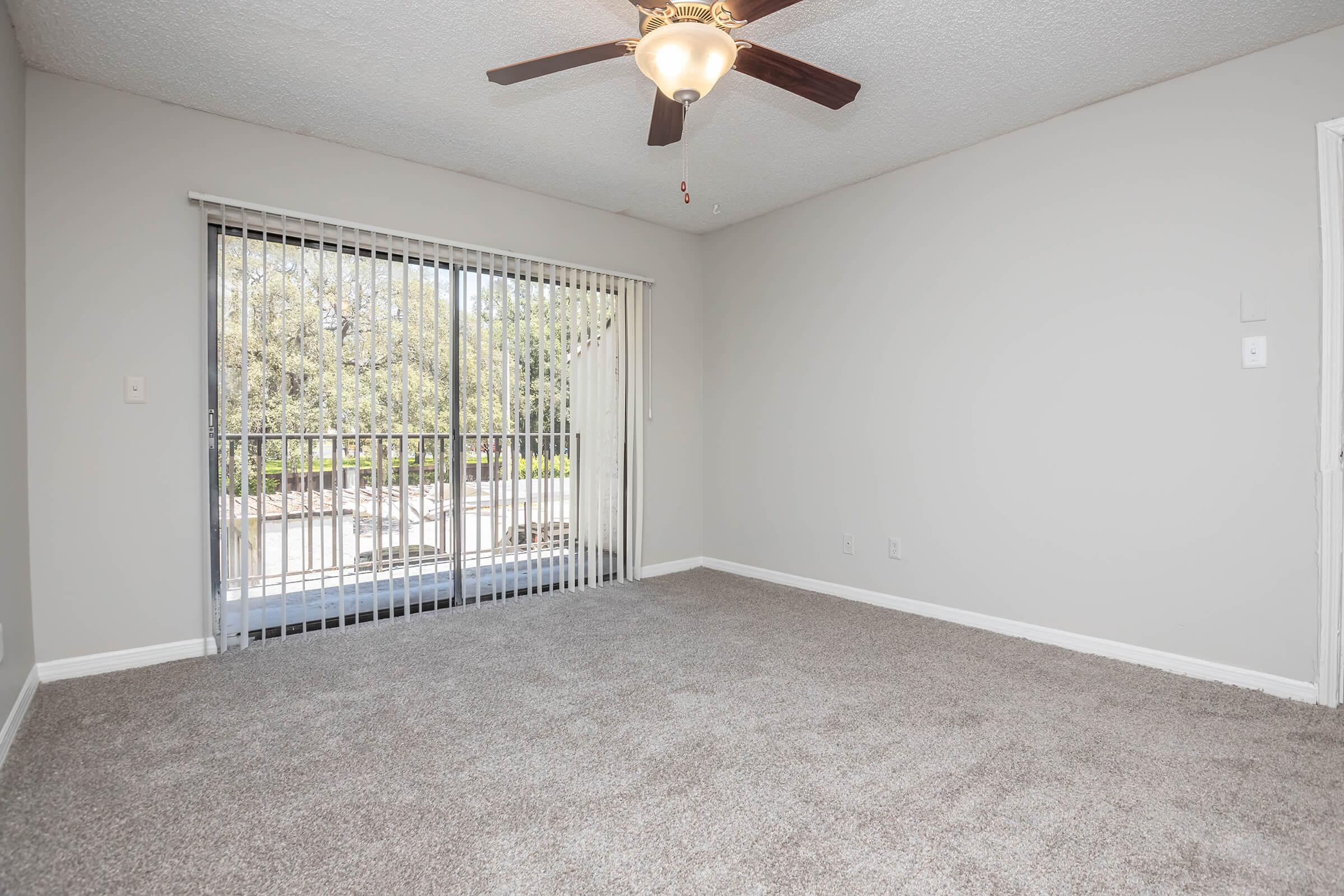 A well-lit living room featuring light gray walls and a ceiling fan. The floor is carpeted in a neutral tone. A glass sliding door leads to a balcony surrounded by trees, providing a serene view. The space is empty, offering a blank canvas for decoration.