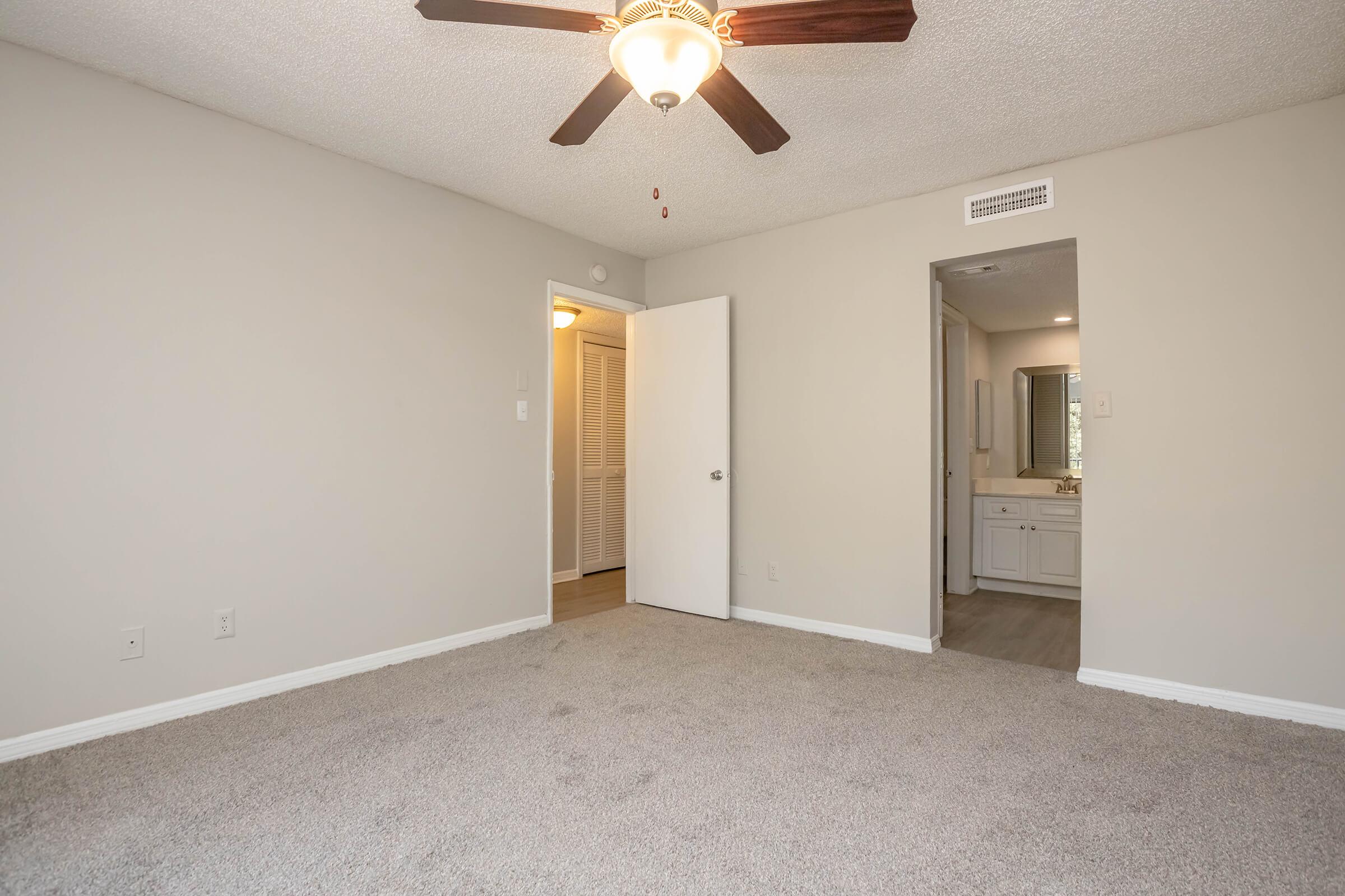 Empty room featuring light-colored walls and carpet, a ceiling fan with wooden blades, a door leading to a bathroom, and another door leading to a closet. Natural light is coming from a window, creating a bright and open atmosphere.
