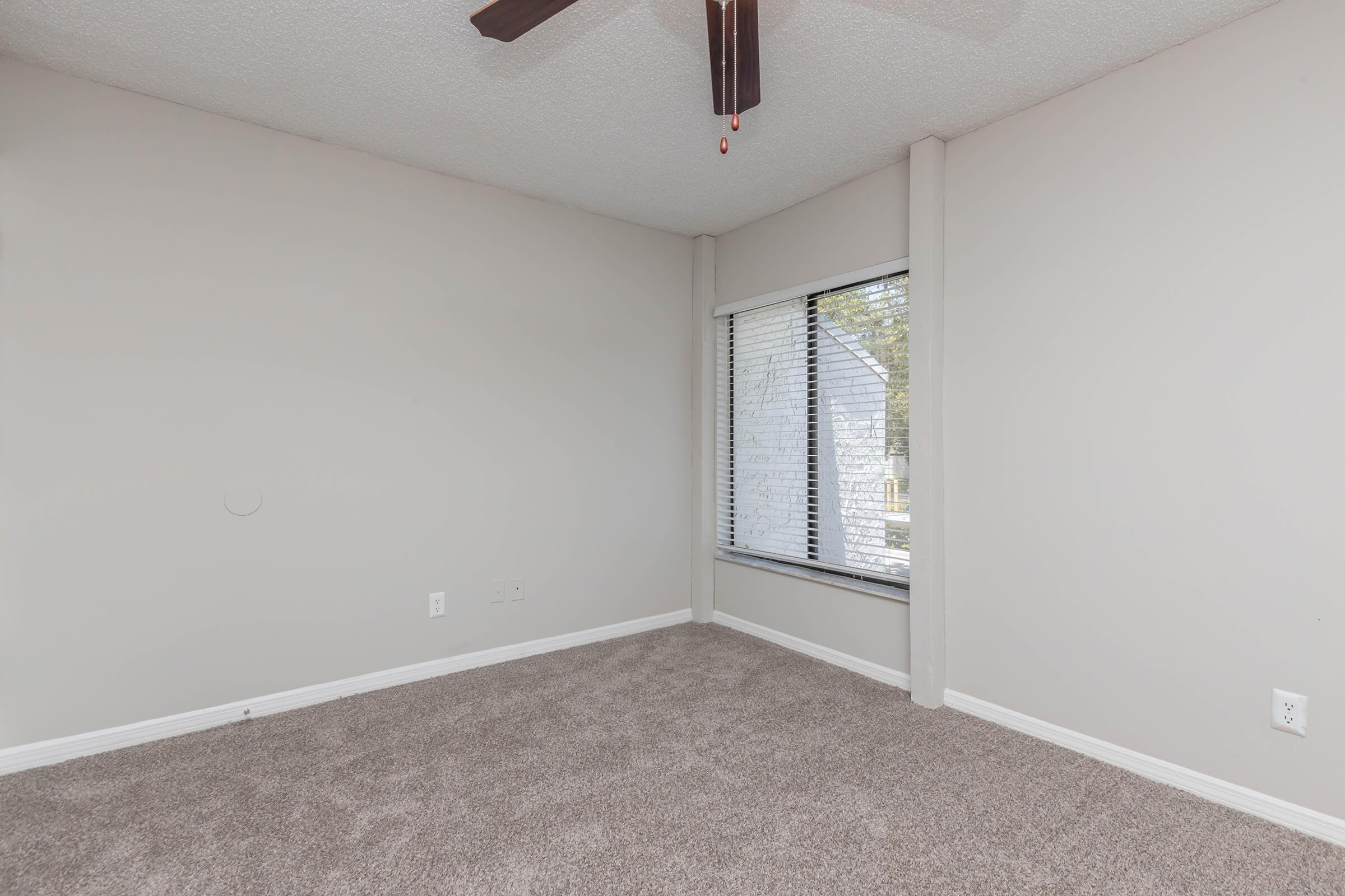 Empty room with beige walls and light brown carpet. A window with vertical blinds lets in natural light, and a ceiling fan hangs above. The space appears clean and well-maintained, ideal for various uses such as a bedroom or office.