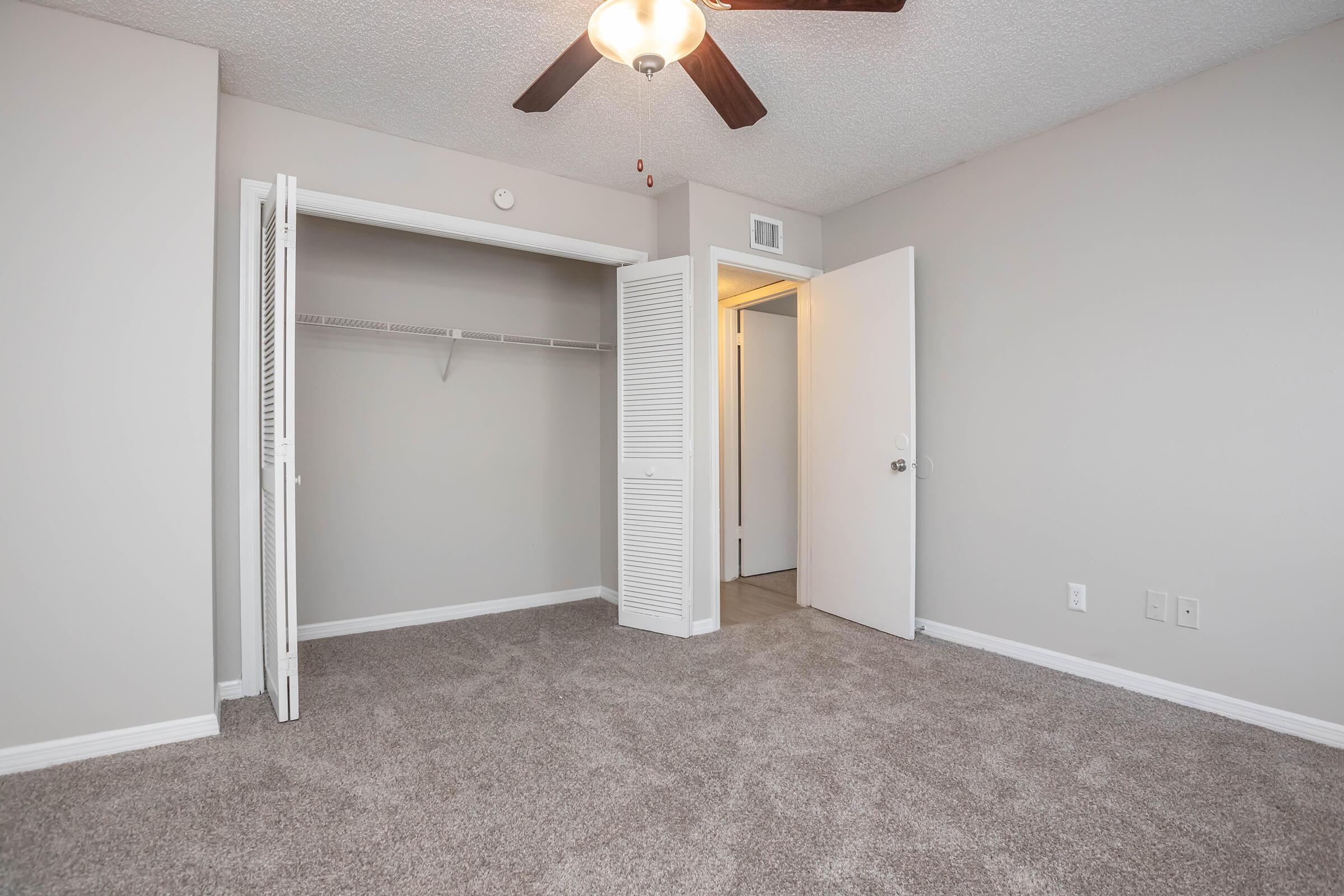 Empty bedroom with light gray walls and plush carpet. A ceiling fan hangs from the ceiling. One closet door is slightly open, revealing a hanging rod. A second door leads to a bathroom or hallway. The overall atmosphere is bright and spacious, with natural light illuminating the room.