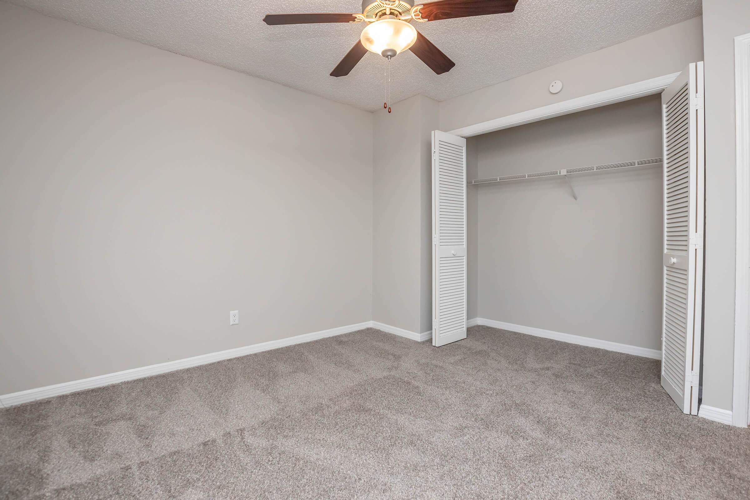Empty room with light gray walls and a ceiling fan. The floor is covered in light-colored carpet. There is a closet with white louvered doors on one side, featuring a hanging rod inside. The room is well-lit, giving a spacious feel.