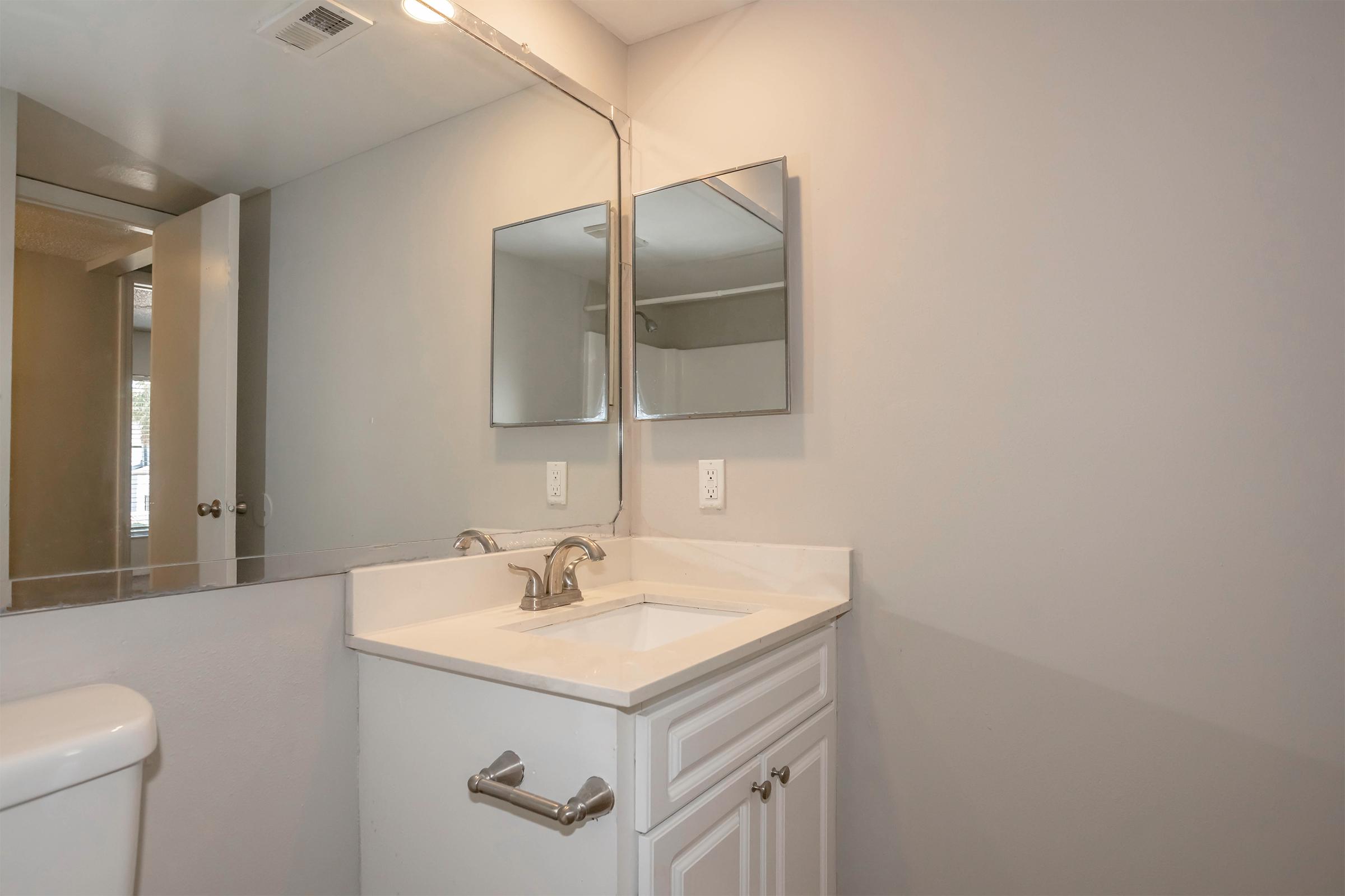 A modern bathroom featuring a white vanity with a sink and two rectangular mirrors above it. The walls are painted a light gray, and there is a white toilet to the left. Natural light is coming from a window, creating a bright and clean atmosphere.