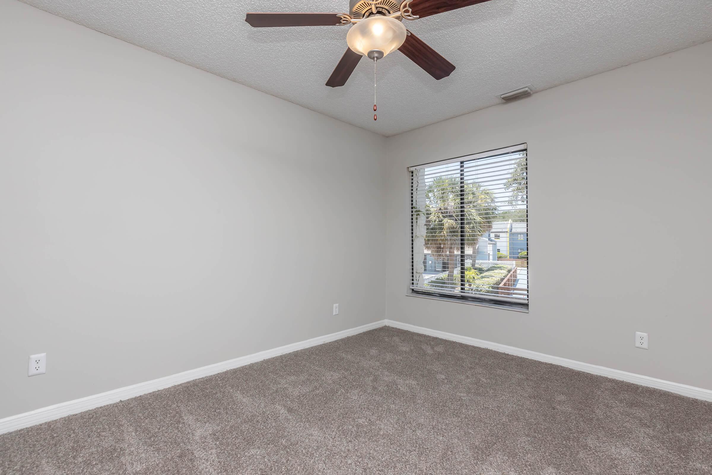 A softly lit, empty bedroom featuring light gray walls, a ceiling fan with wooden blades, and a window with blinds overlooking a green outdoor area. The carpet is plush and beige, providing a warm feel to the space, which is ready for personal décor.