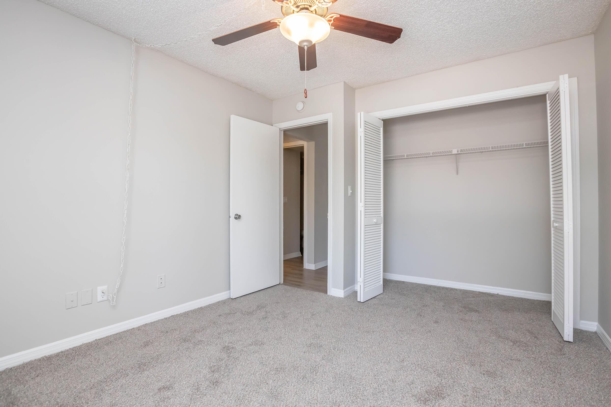 A clean, neutral-colored bedroom featuring light gray walls and plush carpet. The room has a ceiling fan and an open door leading to a closet with bi-fold doors. Natural light enters through the doorway, enhancing the spacious feel.