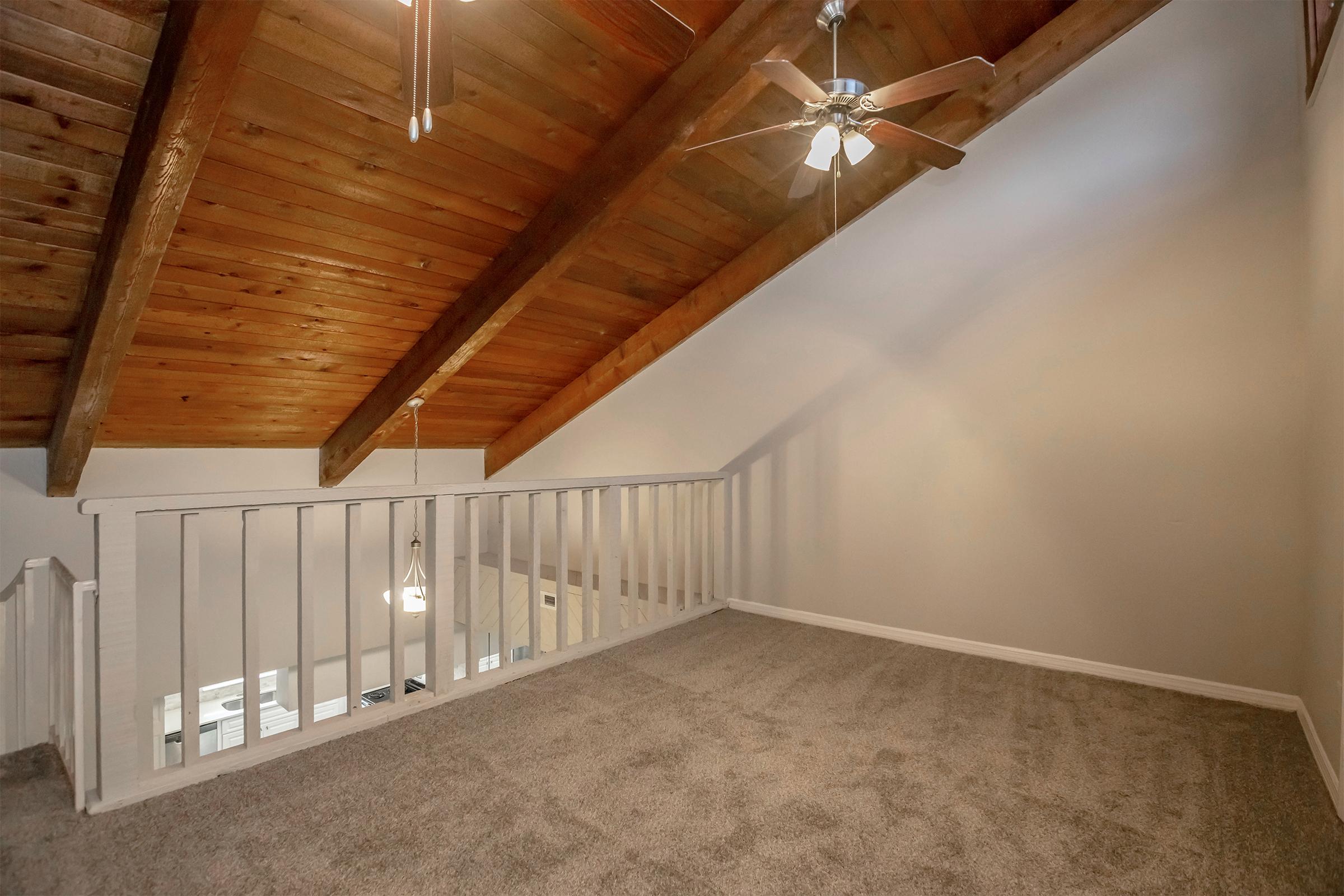 An interior view of a spacious loft area featuring a high, wooden-beamed ceiling and a ceiling fan. The space is carpeted in light gray, with a white railing overlooking the lower floor. Soft, natural light filters in from nearby windows, creating a warm and inviting atmosphere.