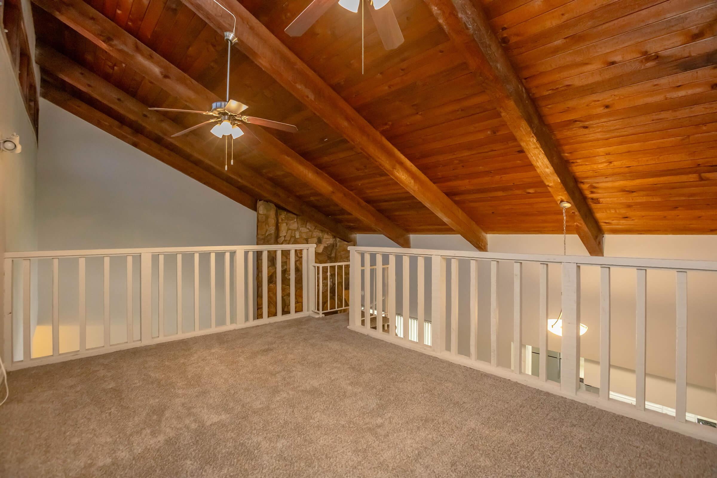 A spacious loft area featuring a high, wooden beamed ceiling and a balcony railing. The floor is carpeted, providing a cozy ambiance, and there are ceiling fans for airflow. The background includes a stone wall and an open layout leading to other parts of the home. Natural light brightens the space.