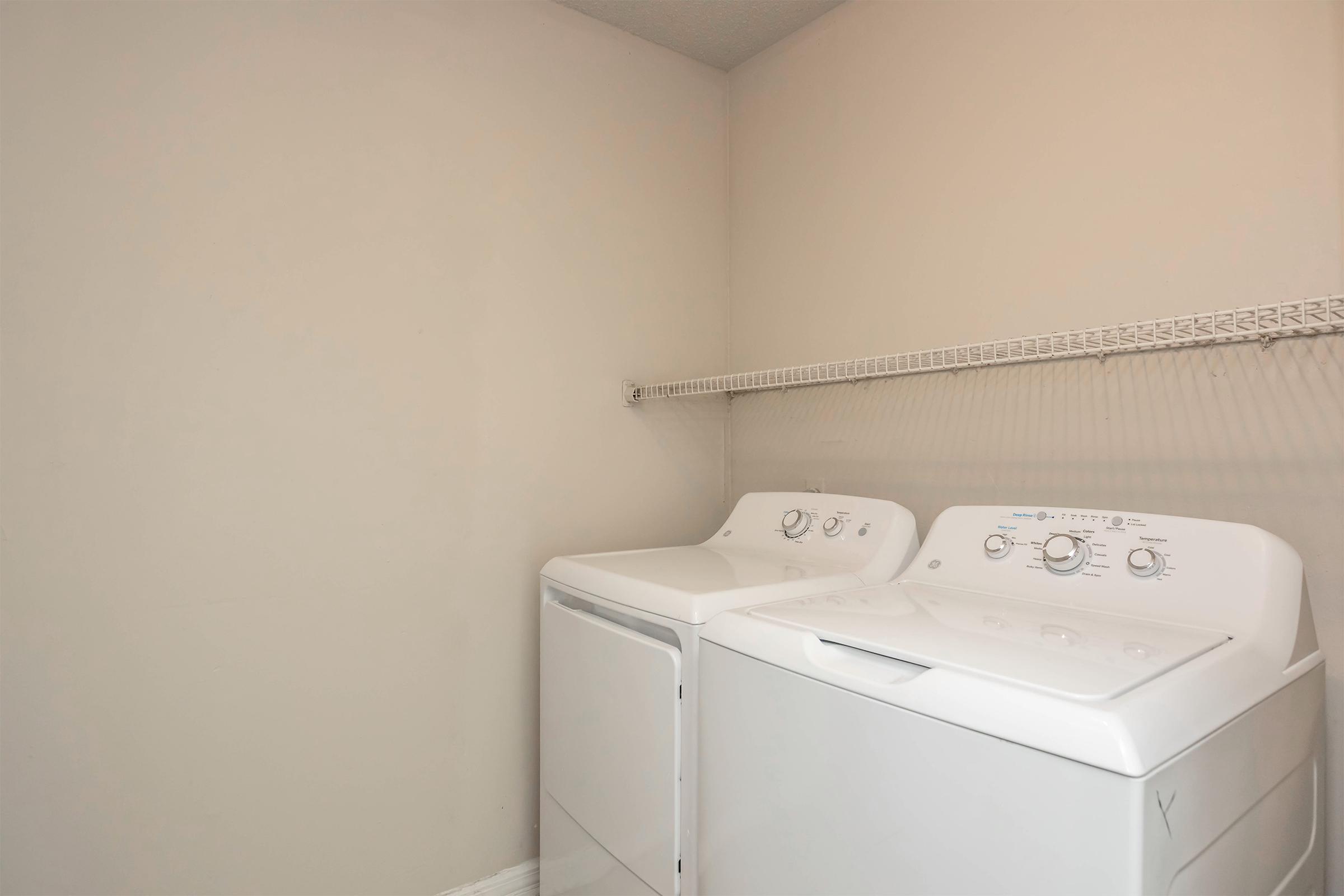 A laundry room with a white washing machine and dryer side by side against a light-colored wall. There is a shelf above them for storage, and the flooring appears to be light-colored as well. The overall space is clean and organized.