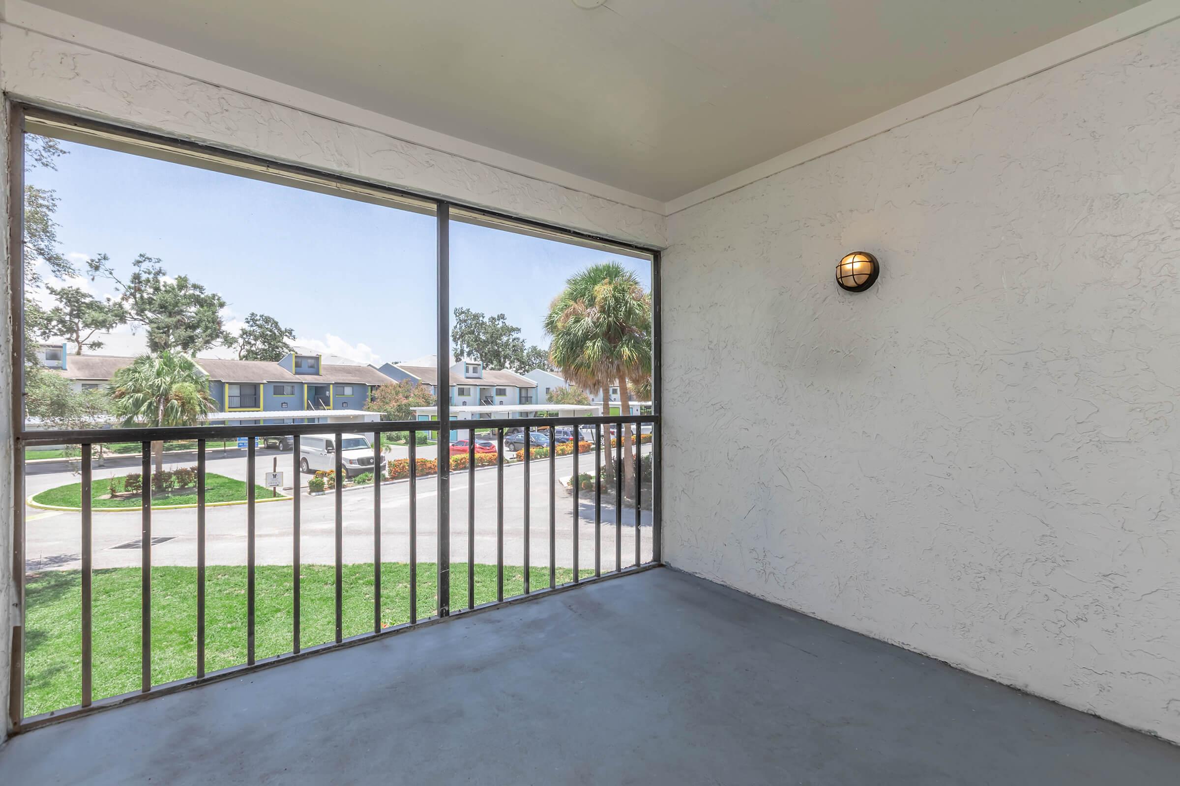 A view from a balcony featuring a concrete floor and a railing, with a clear sky and palm trees visible outside. The background shows a residential area with parked cars. A light fixture is mounted on the wall inside.