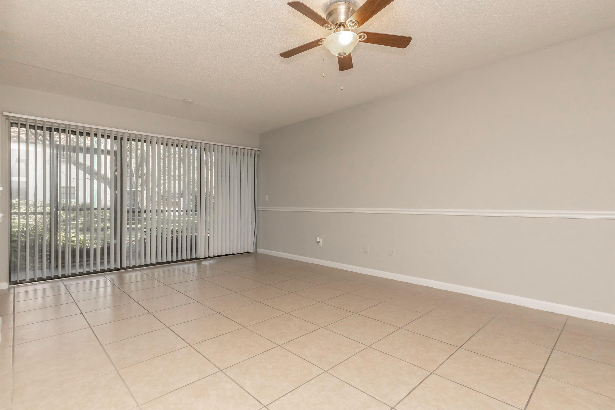 A spacious, empty living room with light-colored tile flooring, a ceiling fan with wooden blades, and a large window covered by vertical blinds that provides natural light. The walls are painted in a soft gray color, enhancing the airy feel of the space.