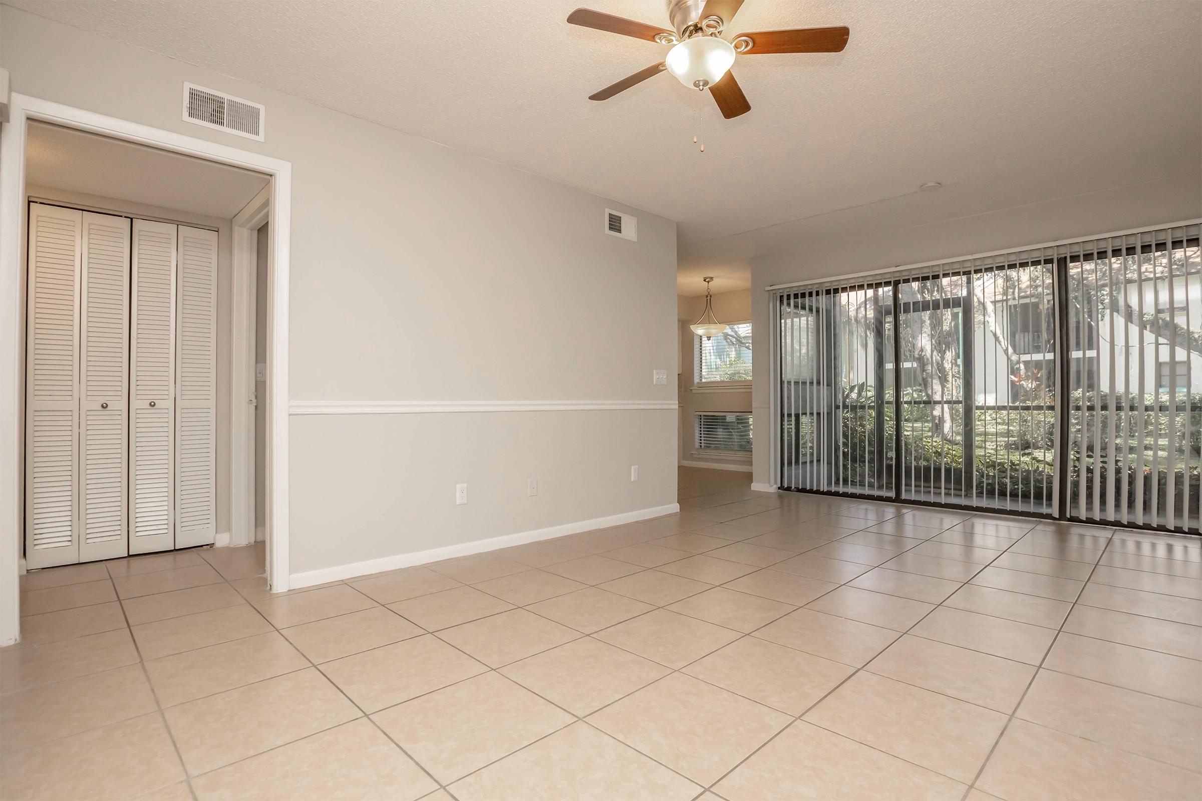 A spacious living area featuring beige tiled flooring, a ceiling fan, and a sliding glass door leading to an outdoor patio. The walls are light grey, with a white chair rail, and there's a closet with double doors on one side. Natural light enters through the patio door, enhancing the room's brightness.