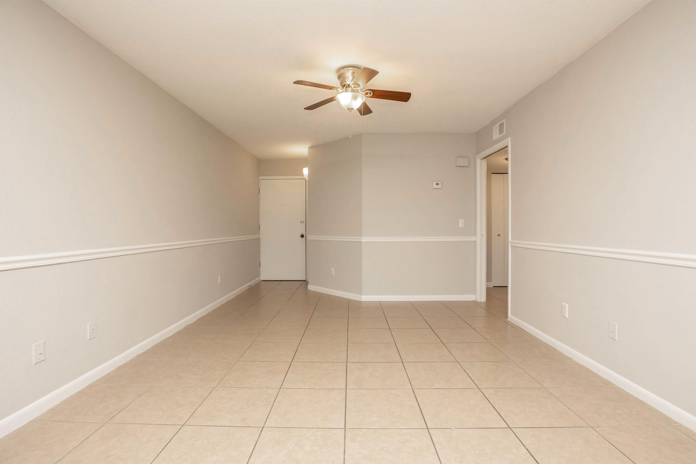 A spacious, empty room with light beige tile flooring. The walls are painted a soft gray, and there's a white chair rail running along the middle. A ceiling fan is mounted in the center, and a doorway leads to a closed door on the right and an entrance door at the far end. The lighting is bright and inviting.