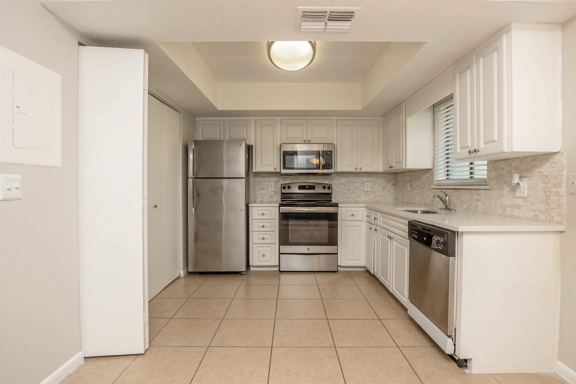 Modern kitchen featuring white cabinetry, stainless steel appliances, a spacious countertop, and tiled flooring. The kitchen includes a refrigerator, oven, microwave, and dishwasher, with a window providing natural light.